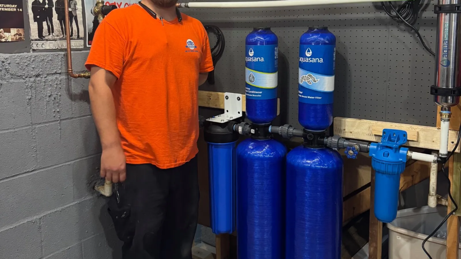 Estes Plumber in an orange uniform stands next to a water filtration system with blue tanks in a workshop setting