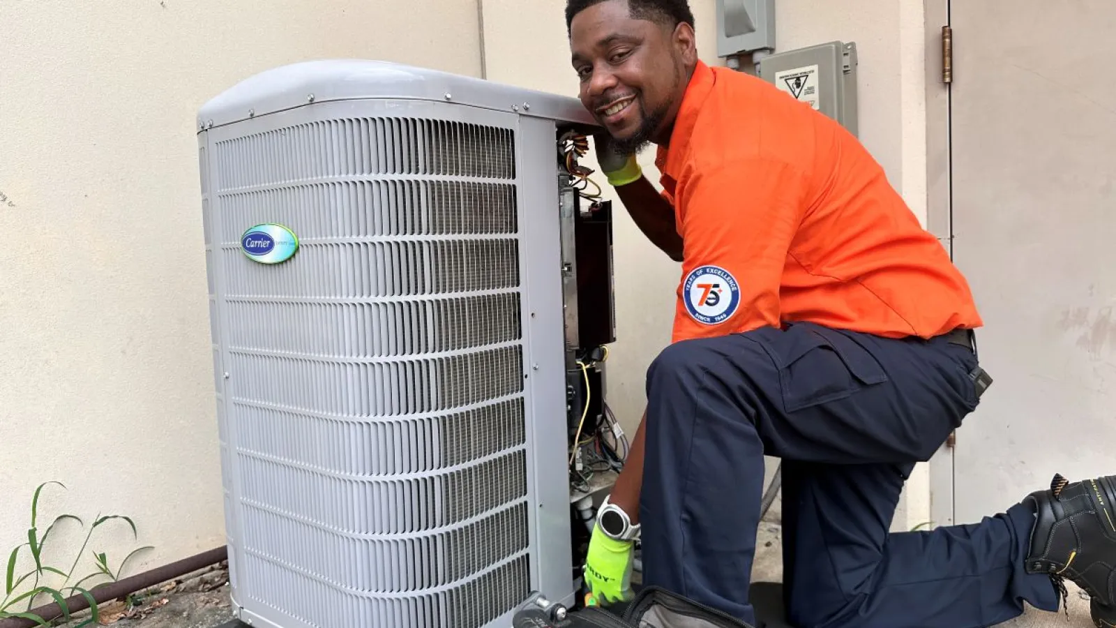 Estes Heating and Air Technician in orange shirt uniform working on outdoor Carrier air conditioning unit with tools on ground in building exterior.