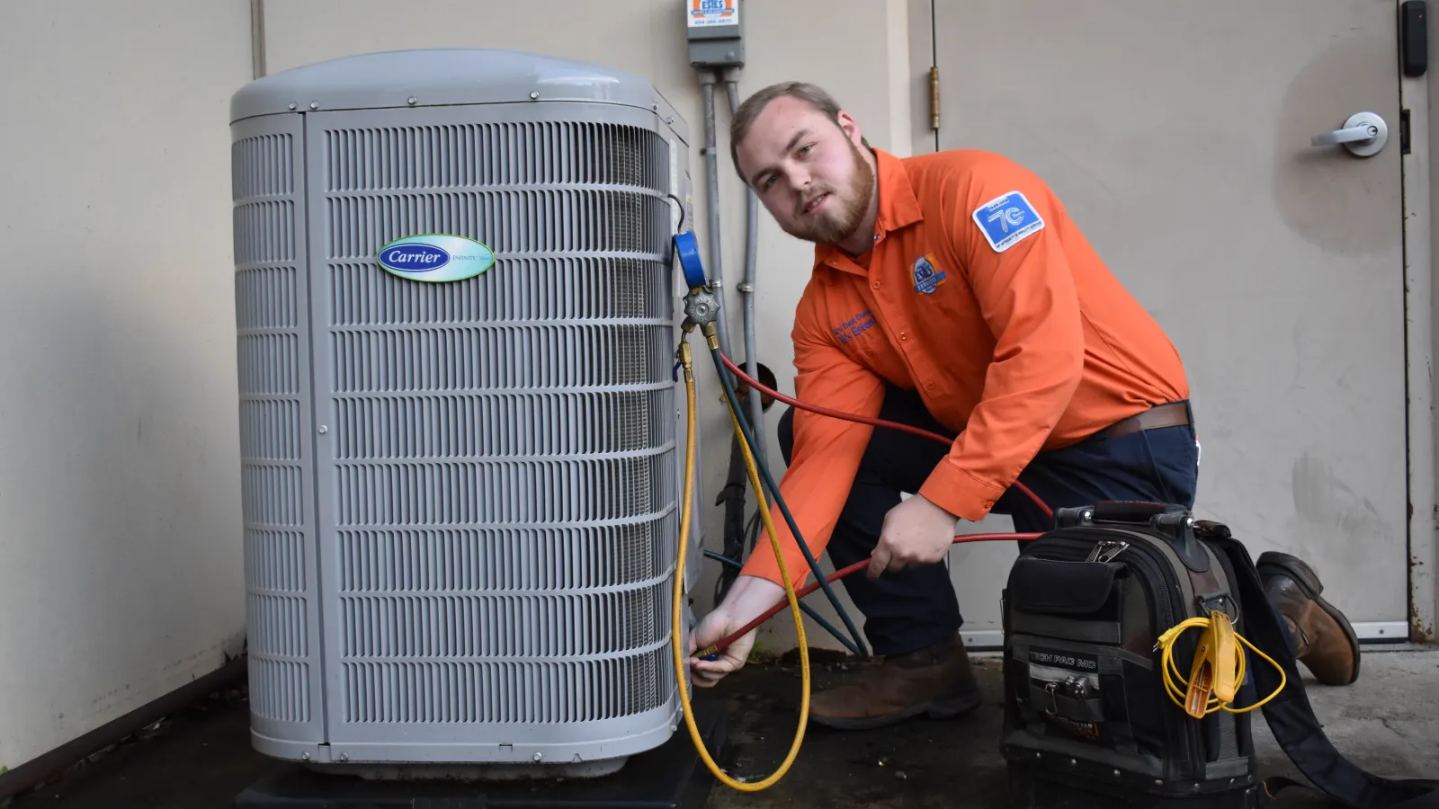 Estes Technician in orange uniform servicing a Carrier HVAC unit with tools and gauges outside a building.