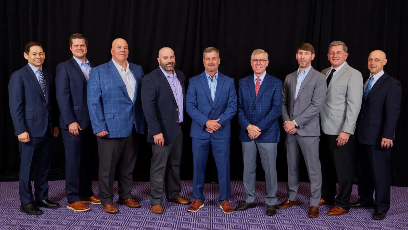 Group of nine professionally dressed men standing in a row against a black backdrop on a patterned carpet.