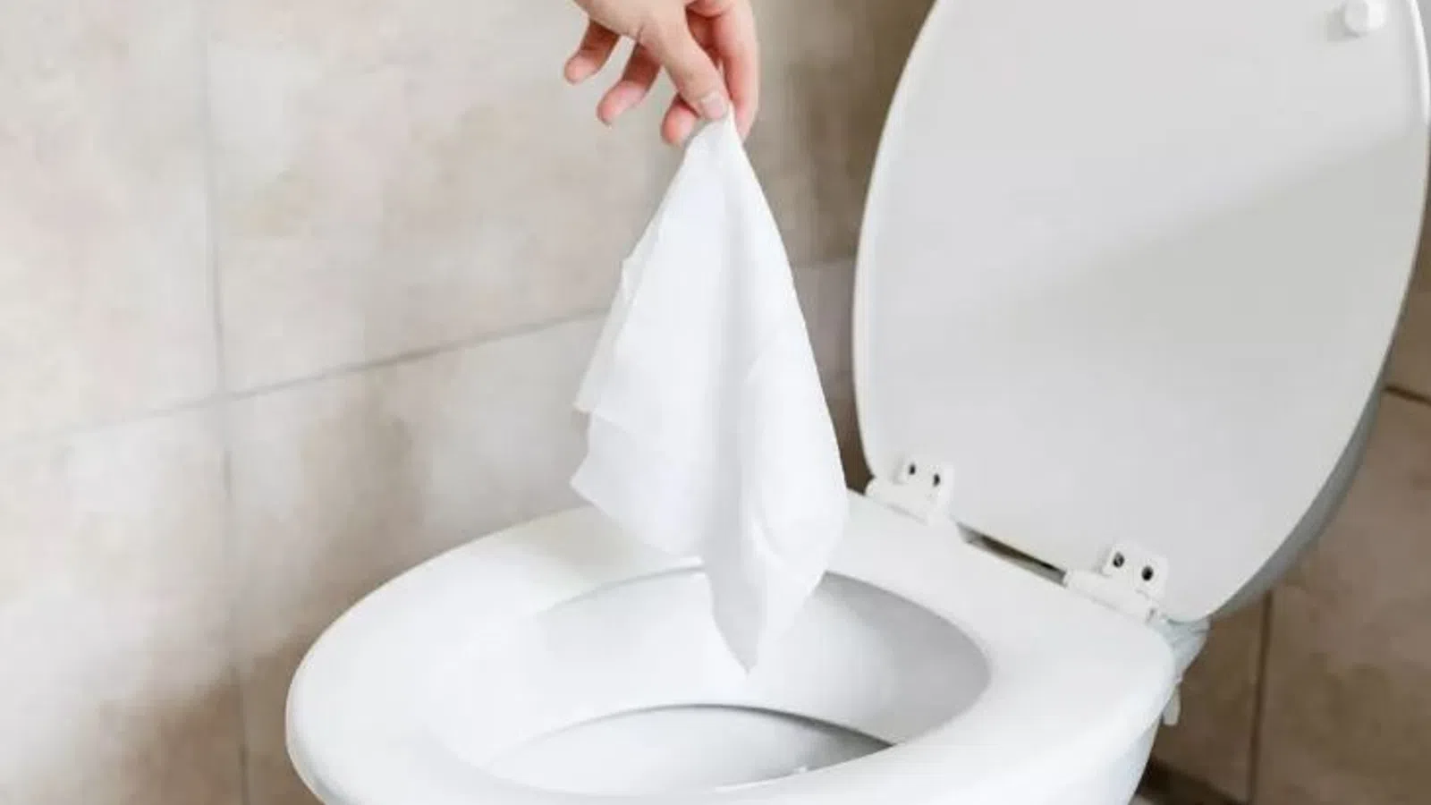Hand holding tissue paper above an open white toilet bowl in a tiled bathroom setting.