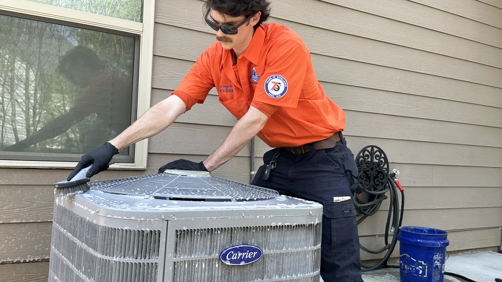 Technician in orange uniform cleaning a Carrier air conditioning unit outside a house on a sunny day.