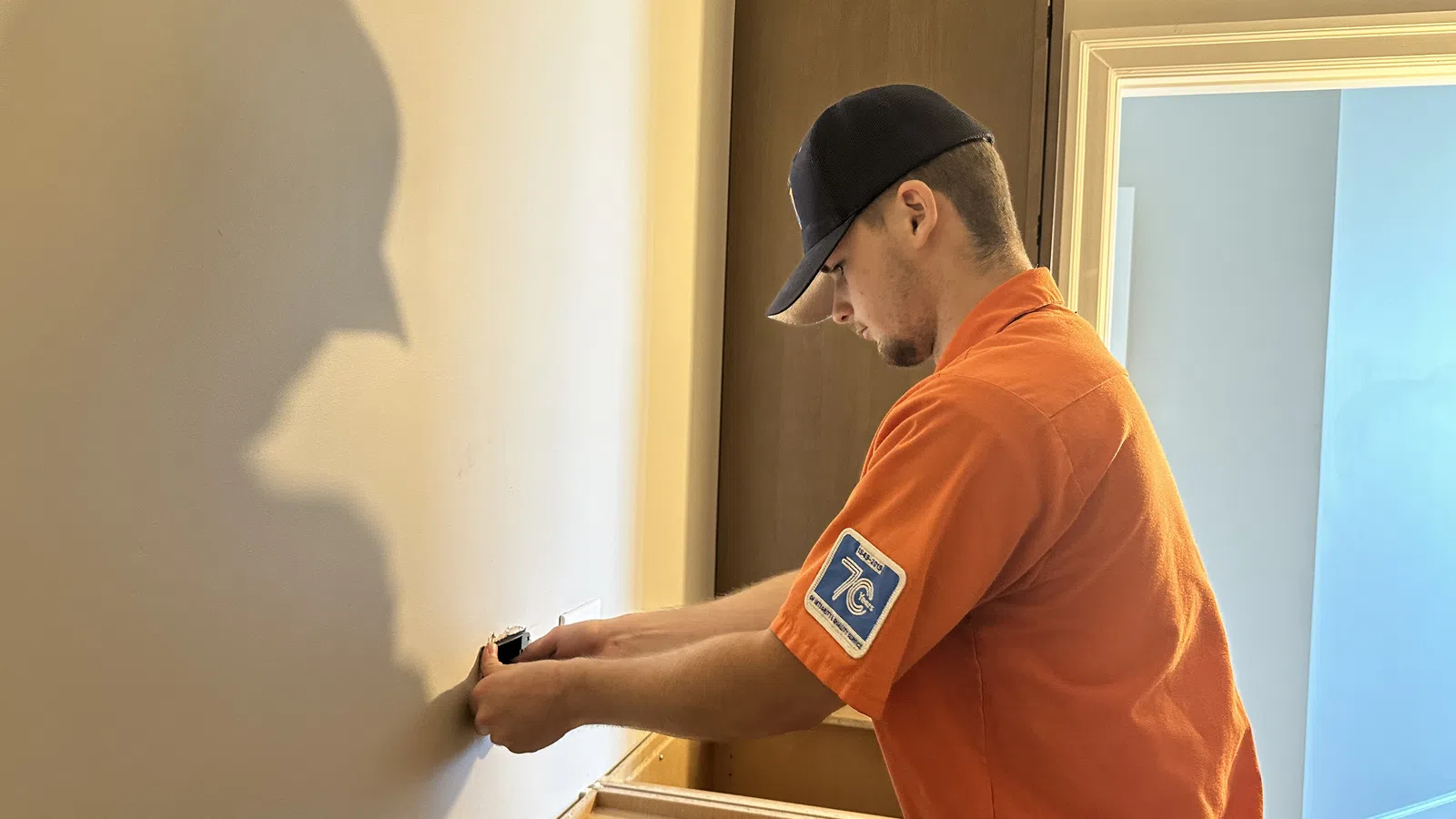 Estes Electriciansin orange shirt and navy cap installing electrical GFCI outlet on a cream wall indoors with open door in background