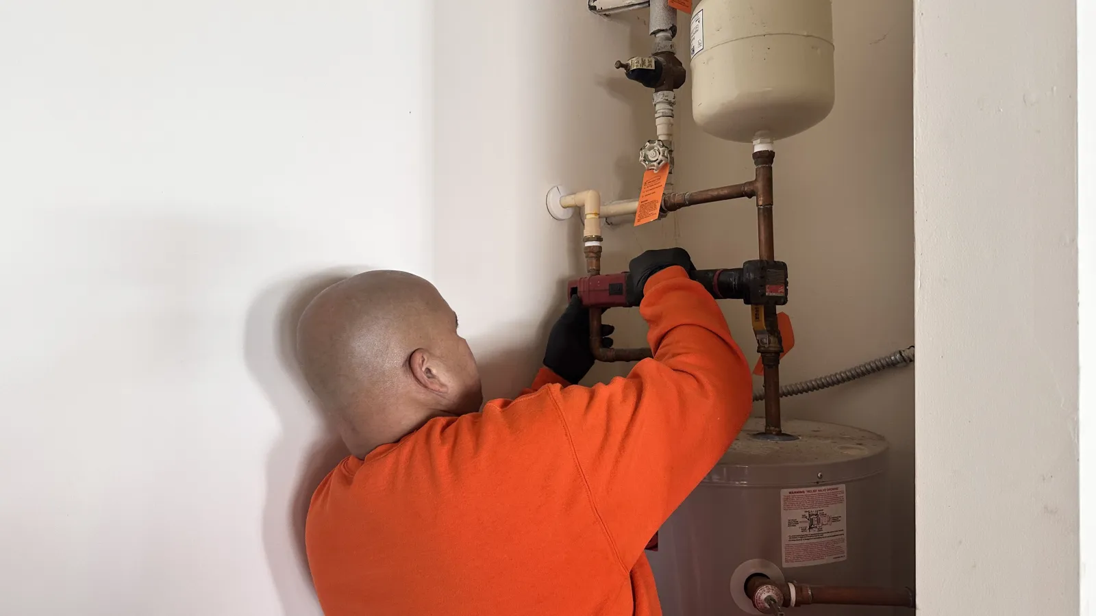 Estes Services Plumber in orange shirt fixing pipes above a water heater in a white utility room corner.