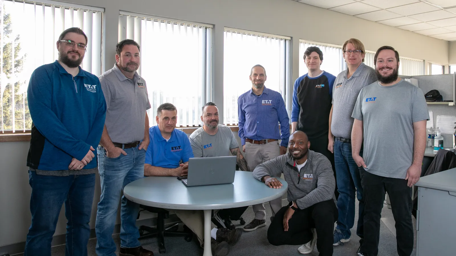 A diverse group of nine professionals posing in an office around a round table with a laptop.