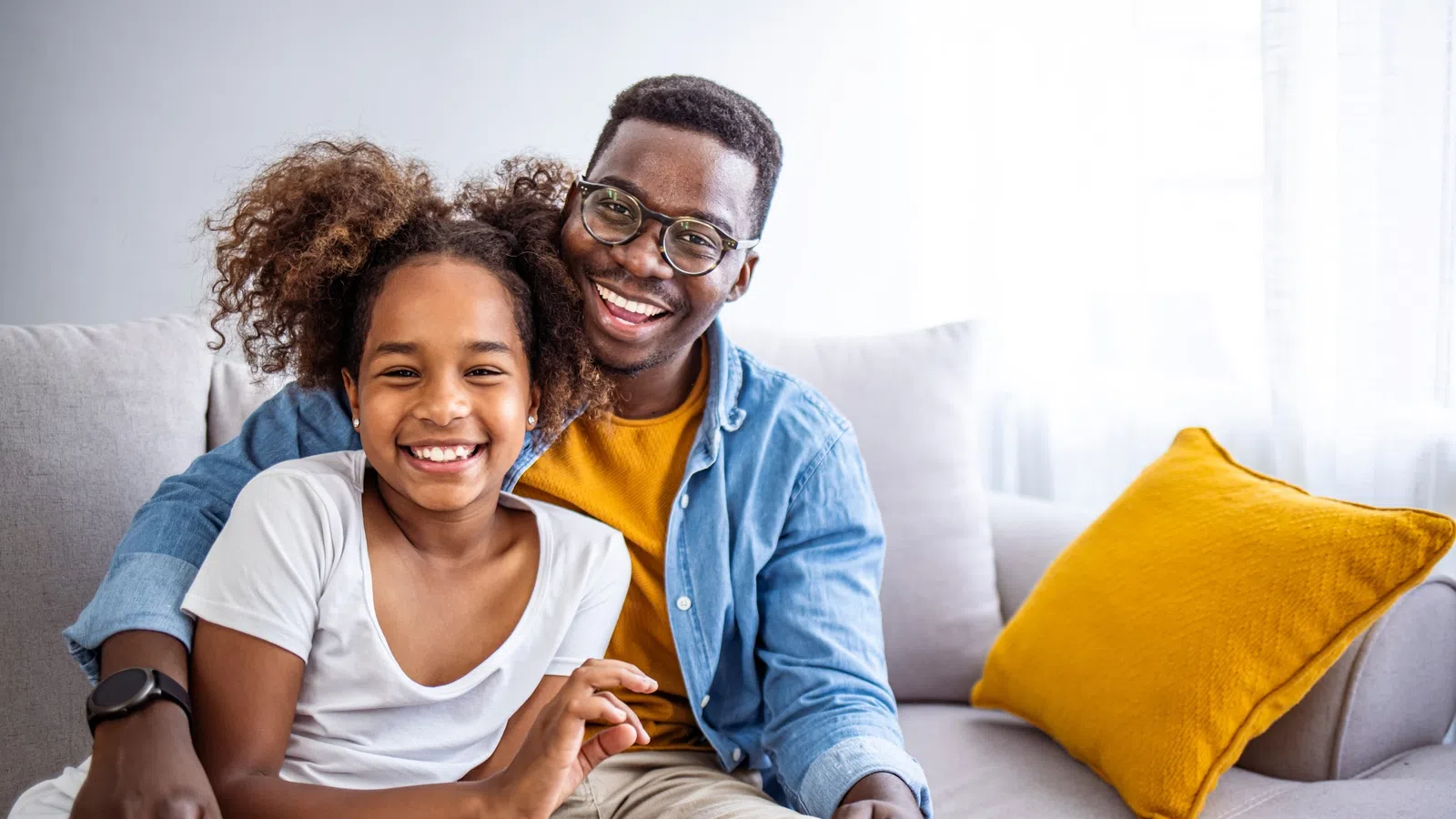 Smiling father and daughter sitting together on a couch in a bright living room with a yellow pillow.