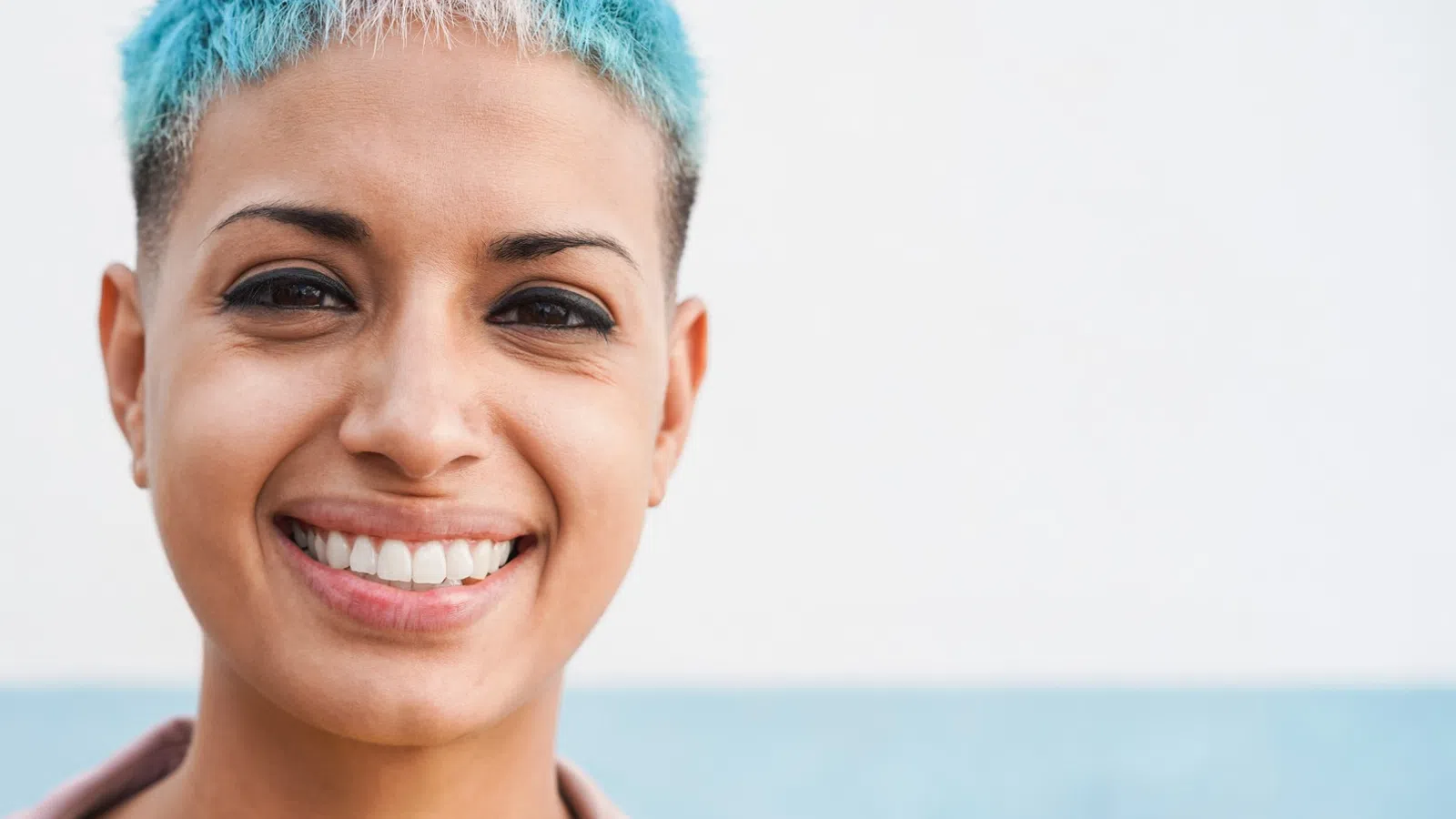 Smiling person with short blue and pink hair wearing a yellow shirt and pink jacket against a light blue background
