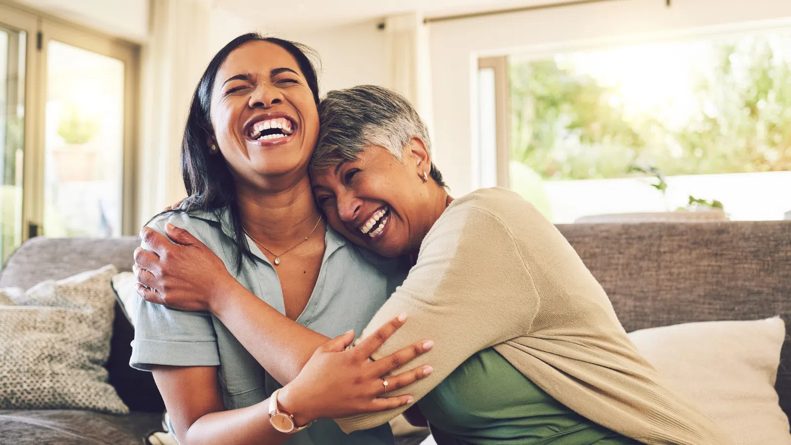 Two women sharing a joyful hug and laughter while sitting on a couch in a bright living room.