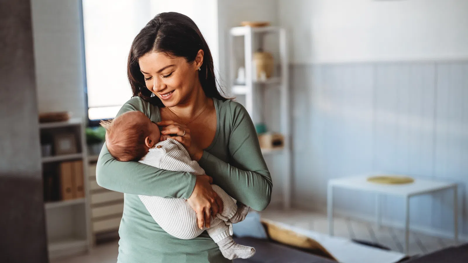 Smiling woman holding and looking lovingly at a sleeping baby in a cozy living room setting.