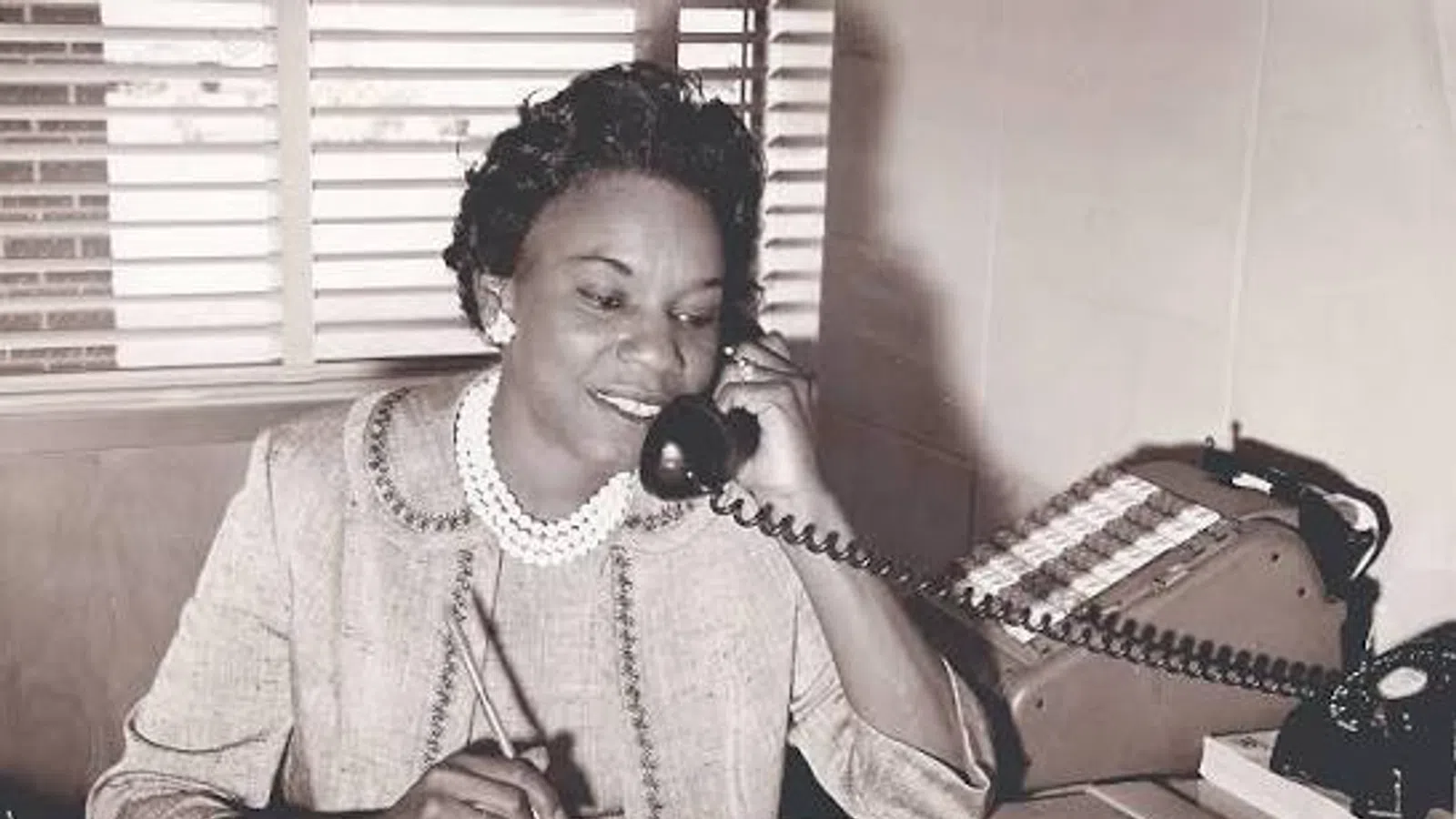Vintage photo of a woman in a pearl necklace talking on a rotary phone while writing at a desk.