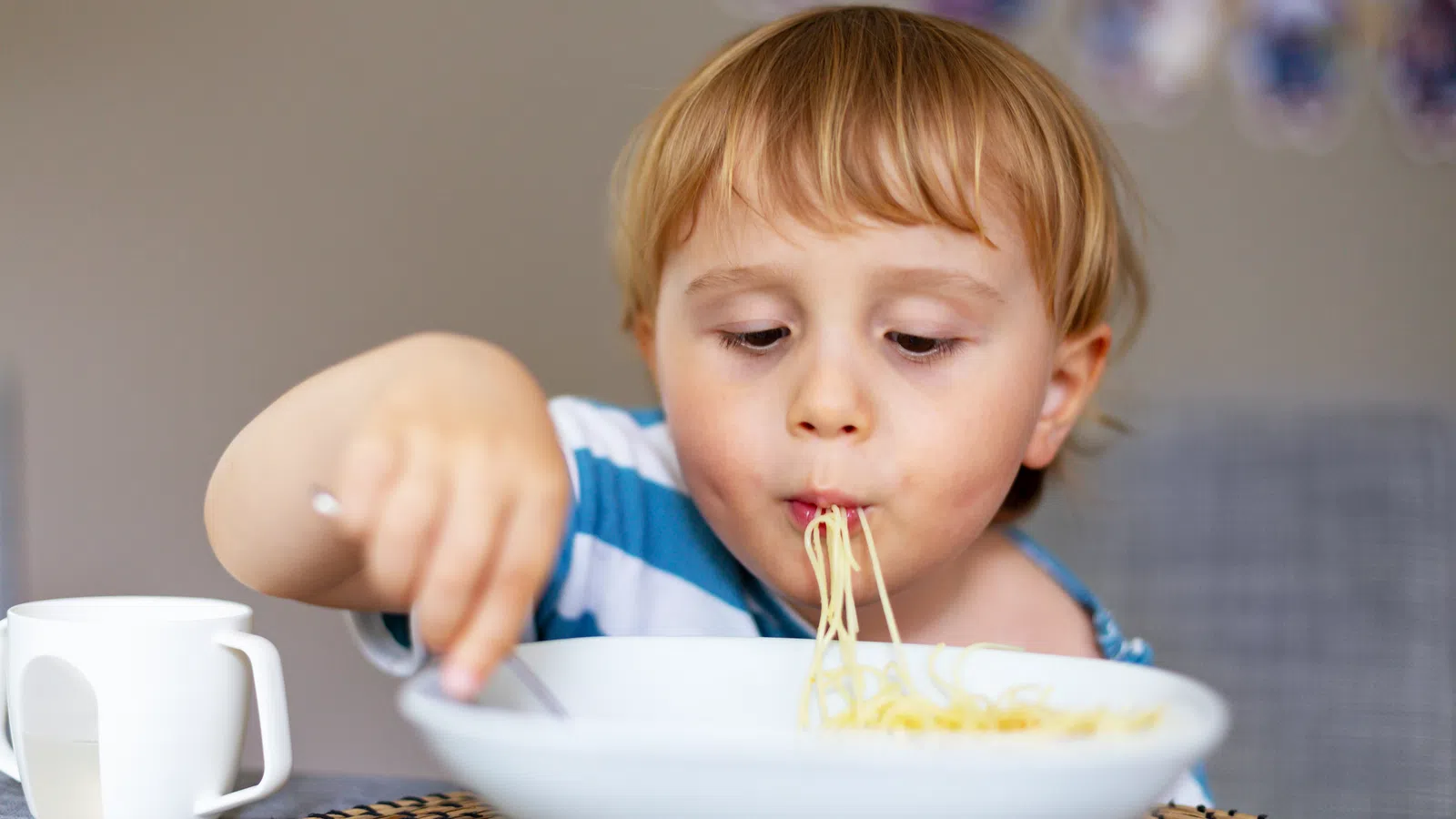 Young child eating spaghetti from a large white bowl at a dining table, focused on enjoying the meal.