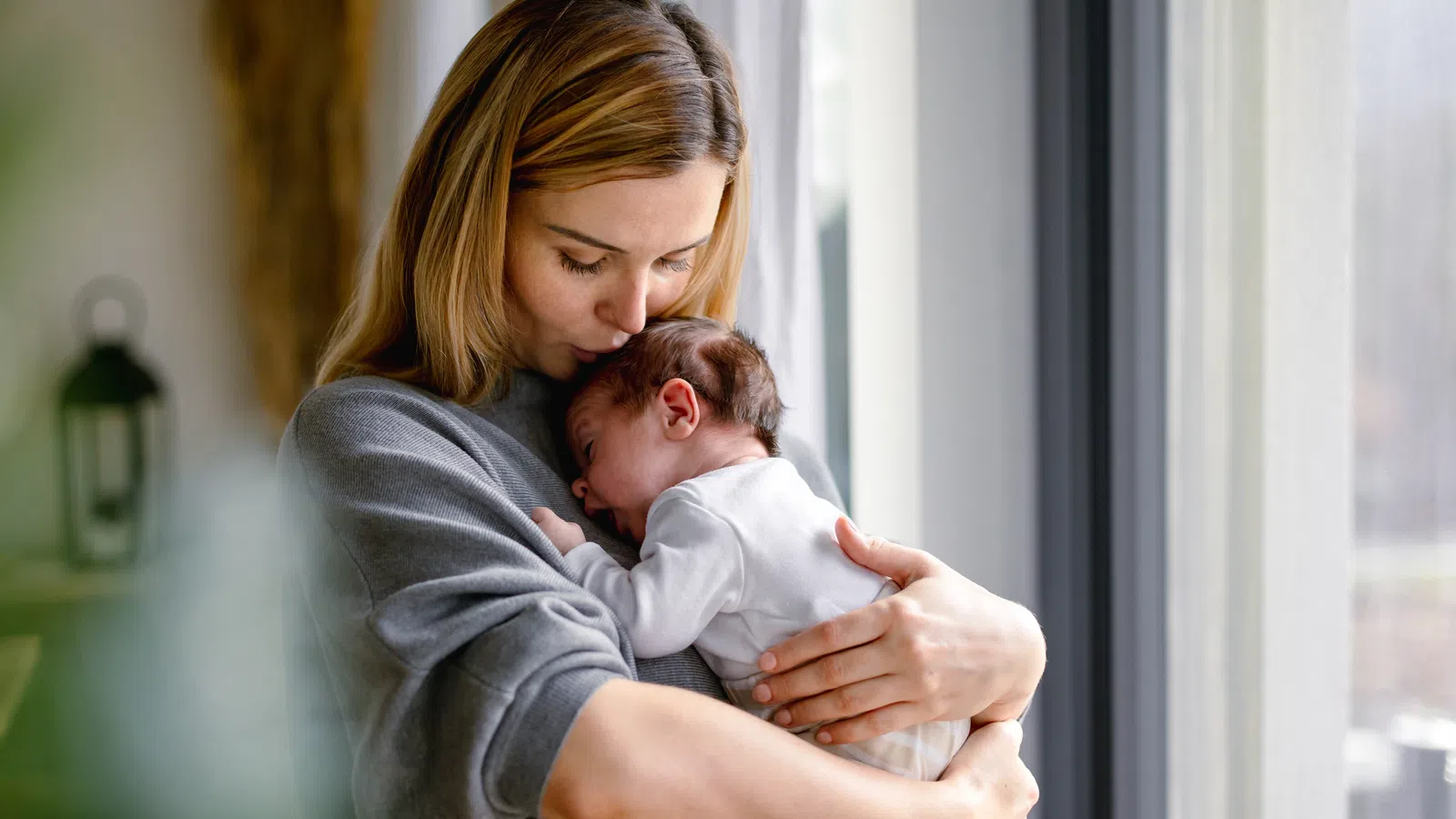 Mother tenderly holding and kissing her newborn baby near a window in a cozy home setting.