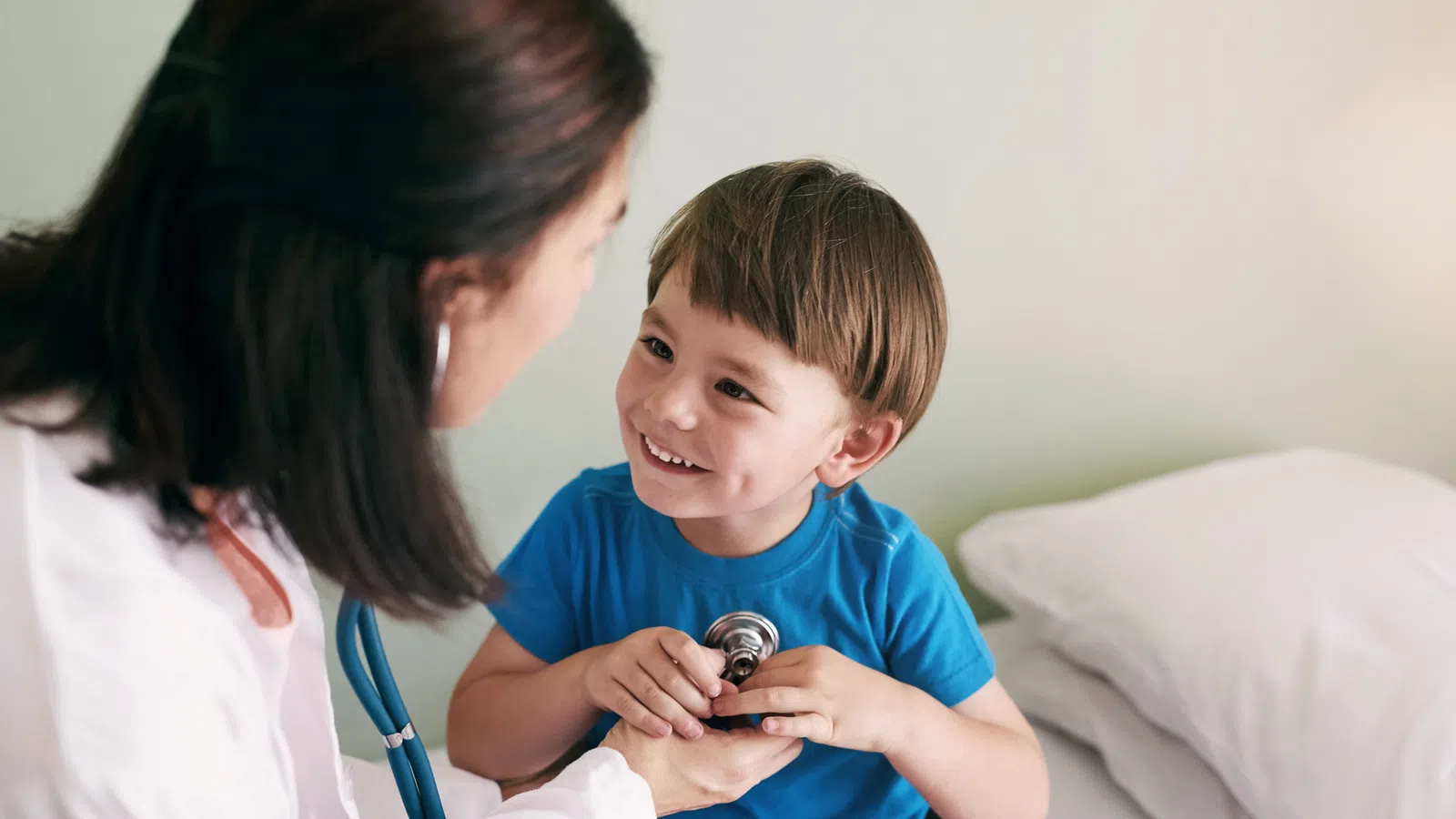 Young boy smiling and holding a stethoscope while sitting on a medical examination bed with a female doctor.