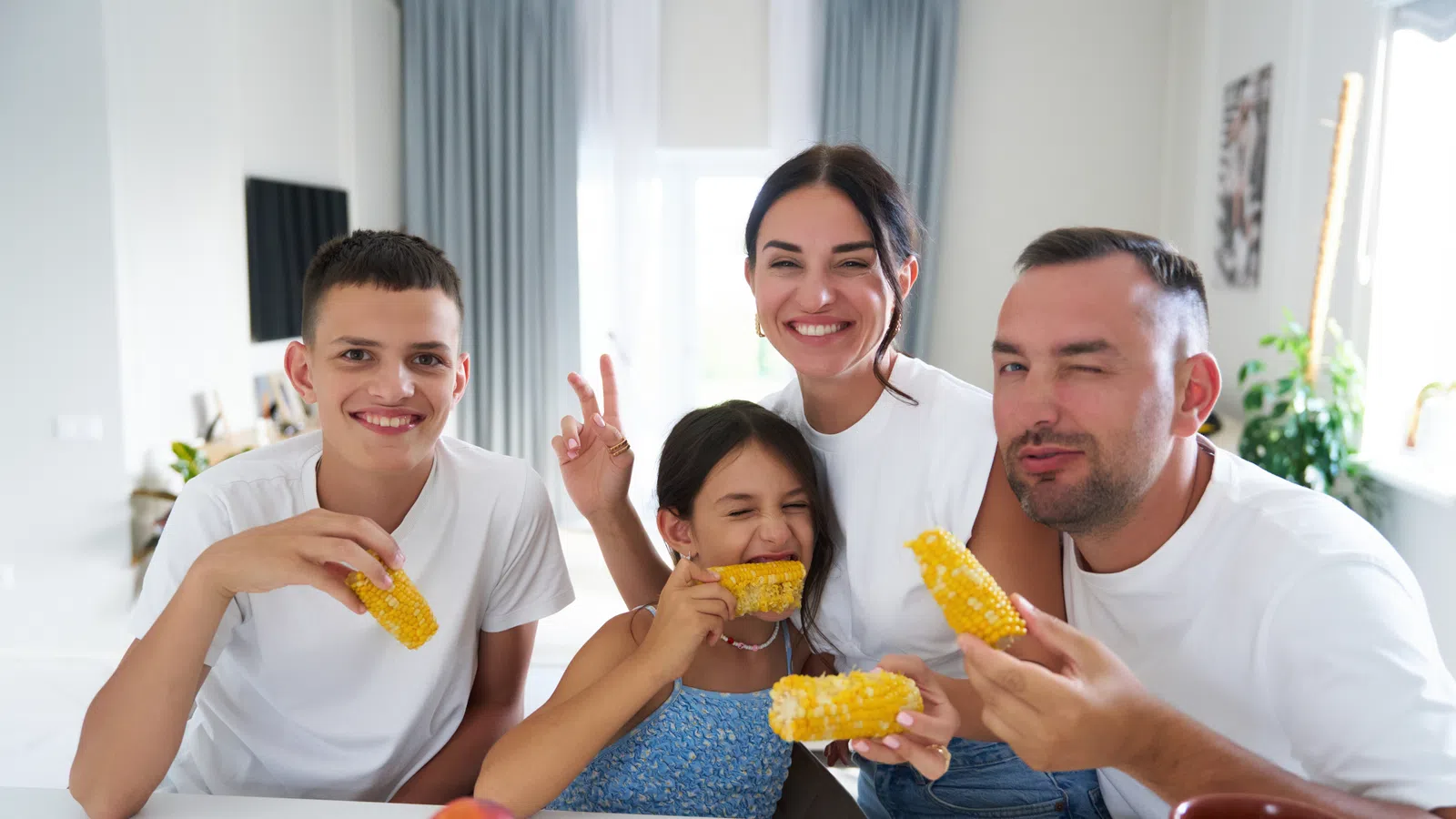 Happy family of four eating corn on the cob together in a bright kitchen with casual white shirts.