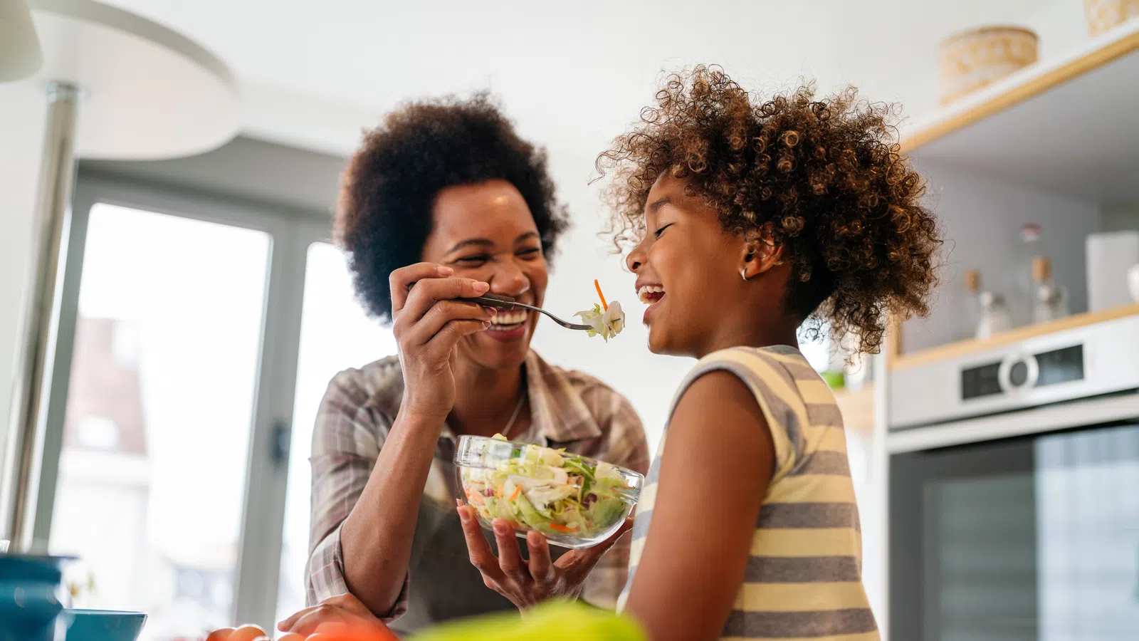 Mother feeding daughter fresh salad in bright kitchen with smiles and playful interaction.