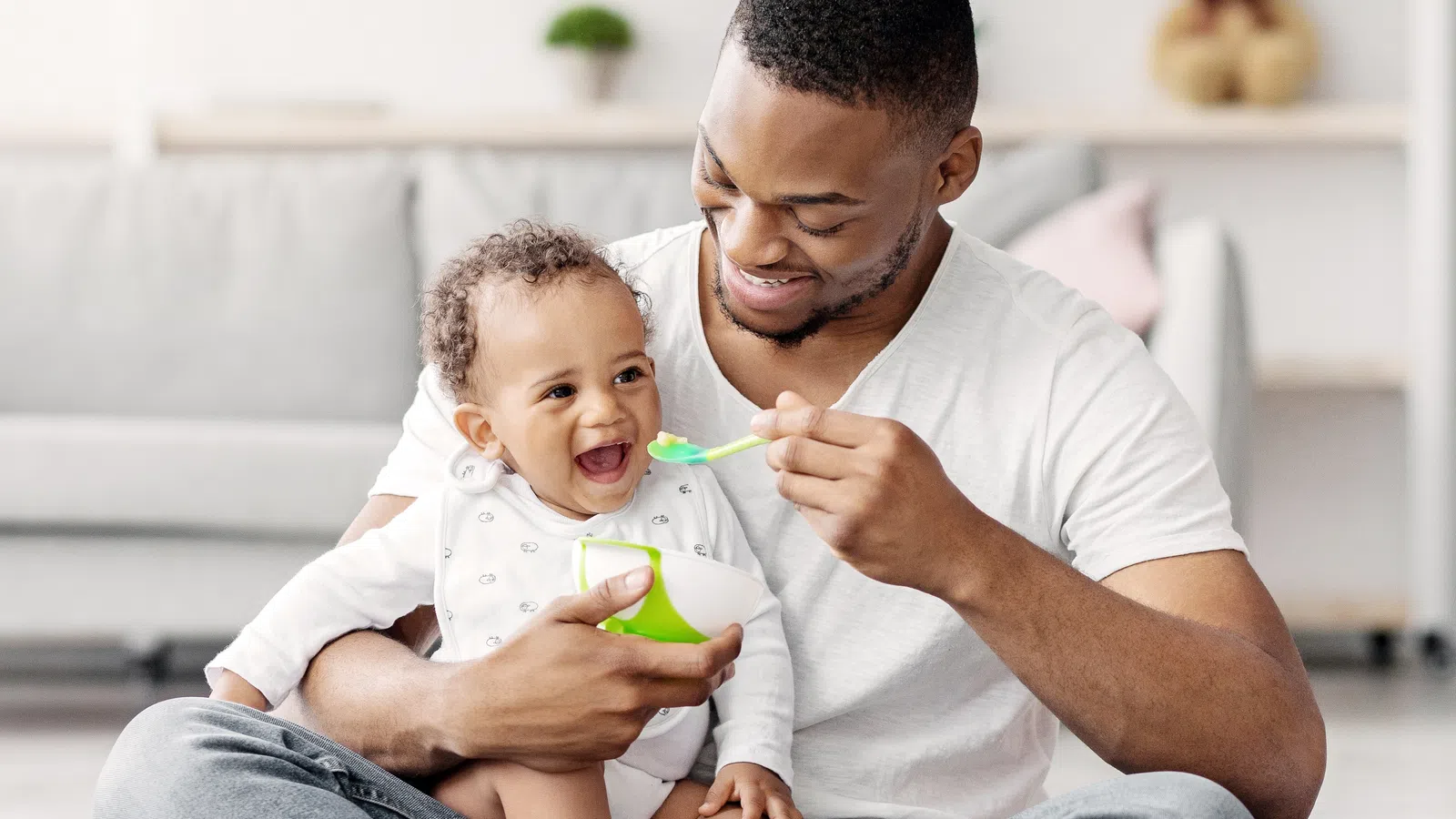 Father feeding baby with spoon and bowl, both smiling and sitting in a bright living room.