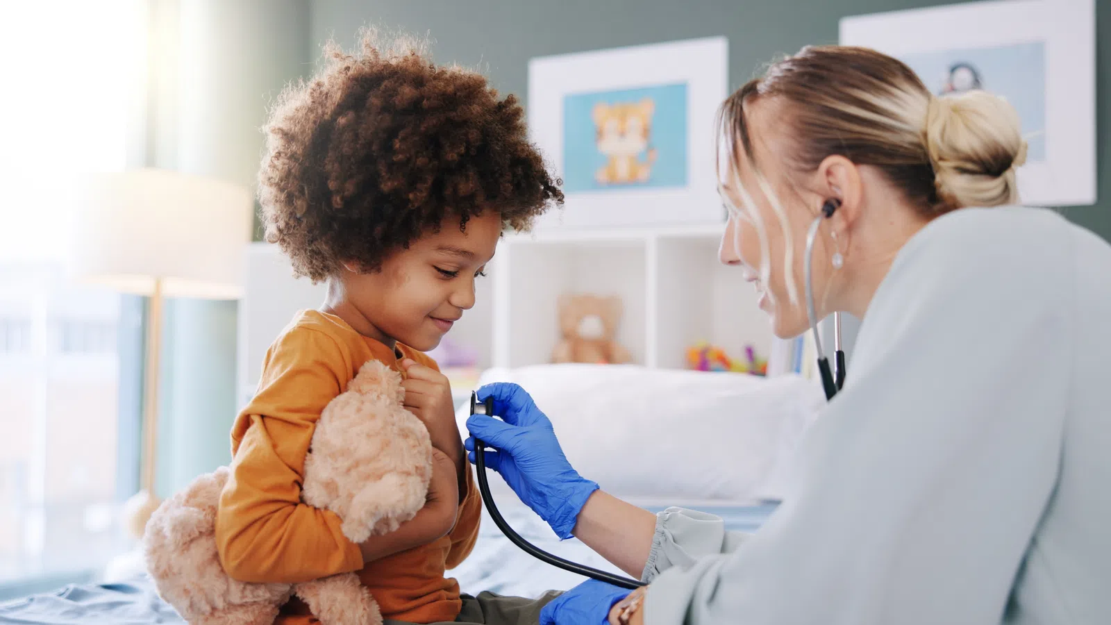 Child holding teddy bear while doctor with gloves listens to heart using stethoscope in bright clinic room