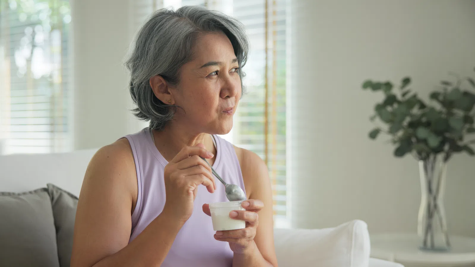 Mature woman in purple sleeveless top eating yogurt with spoon while sitting on a white couch indoors.