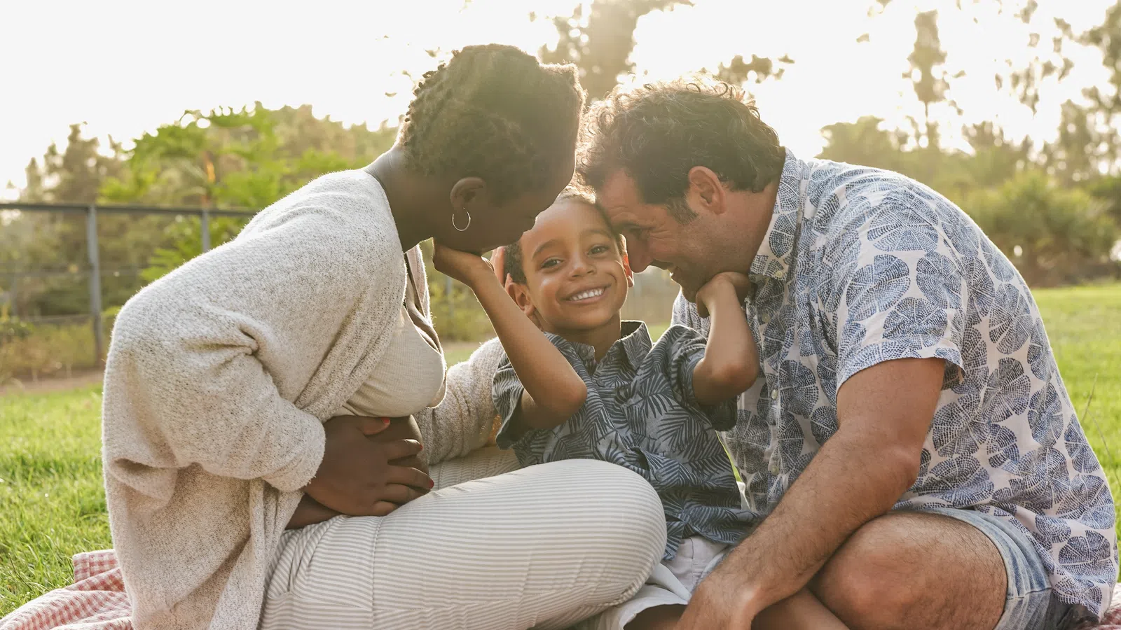 Happy family of three sitting on blanket in park enjoying sunny day with smiles and affection.