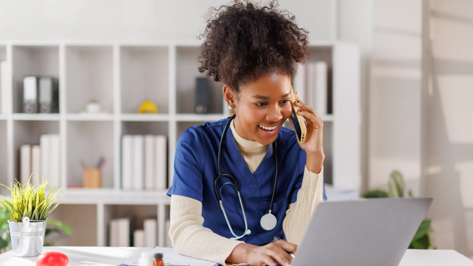 Smiling nurse in blue scrubs talking on phone while using laptop at bright office desk with medical items.