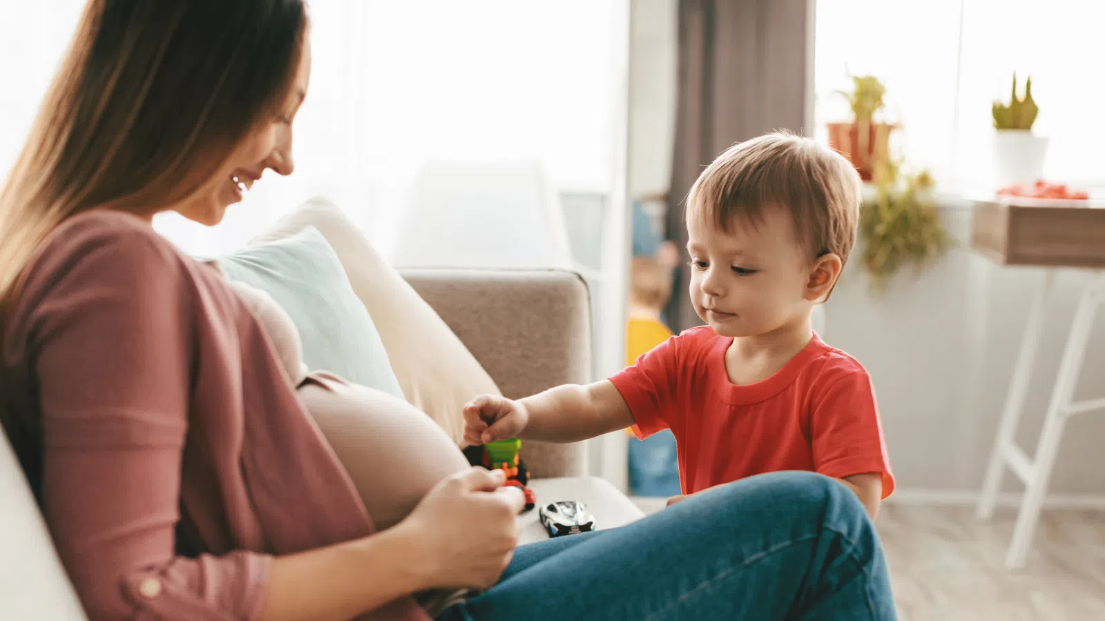 Pregnant woman sitting on a couch smiling as her young son plays with toys nearby in a cozy living room.