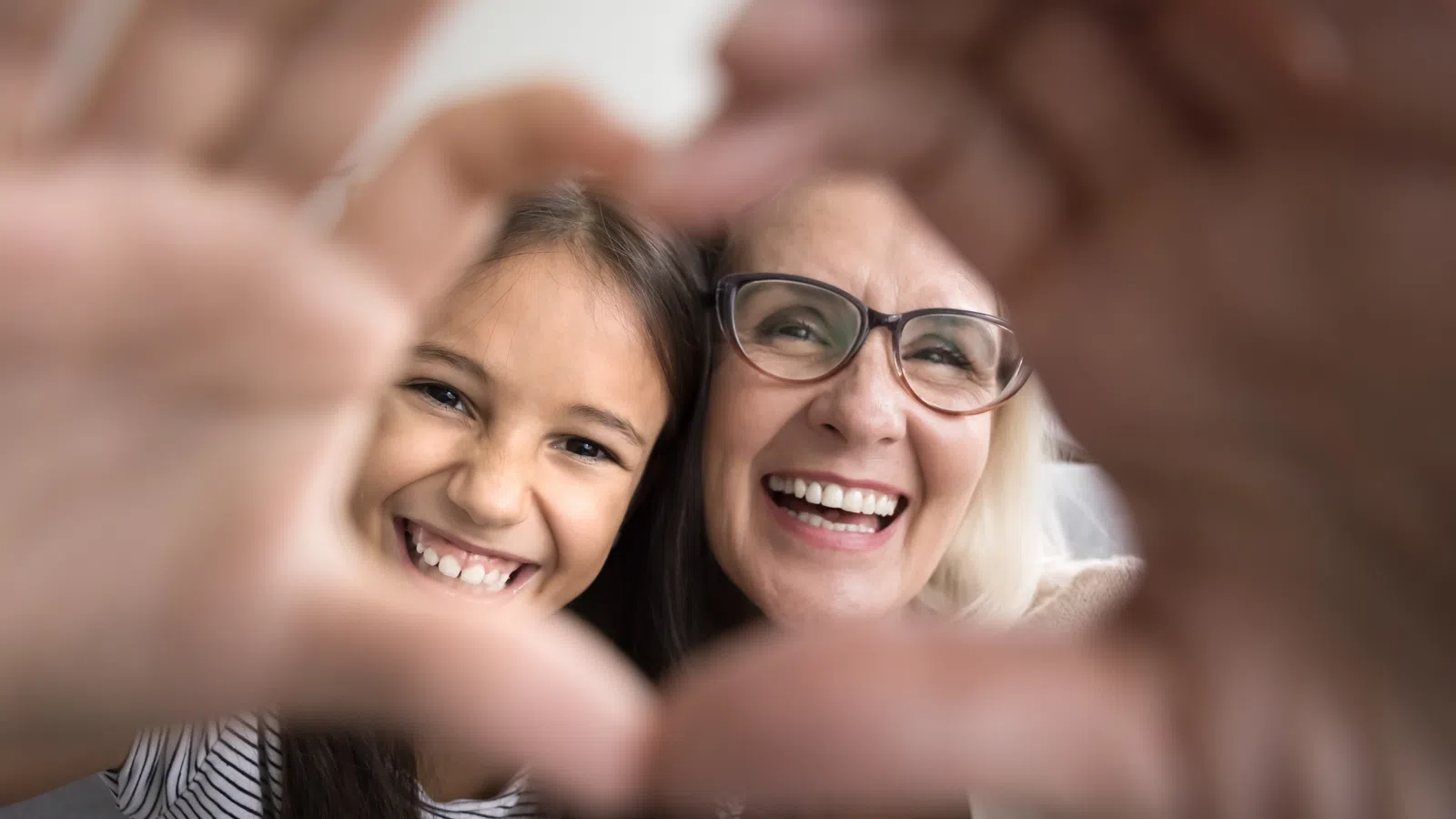 Smiling grandmother and granddaughter forming a heart shape with their hands and looking through it.