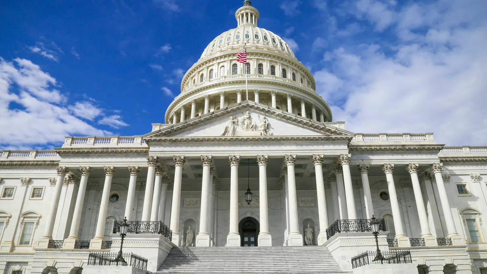Front view of the United States Capitol building under a partly cloudy blue sky with an American flag flying.