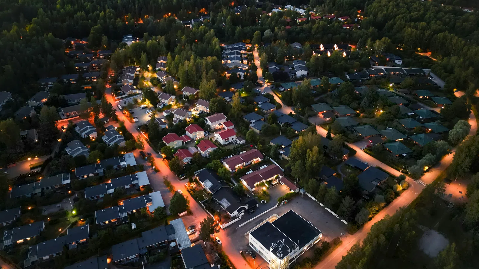 Aerial night view of a suburban neighborhood with illuminated streets and dense tree coverage surrounding homes.
