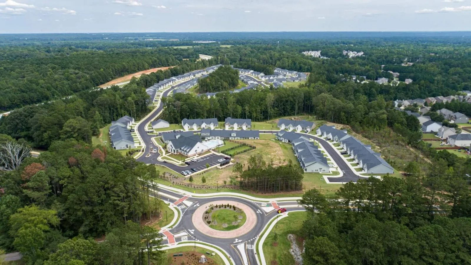 Aerial view of a residential neighborhood with multiple houses surrounding a roundabout amid dense green forest areas.