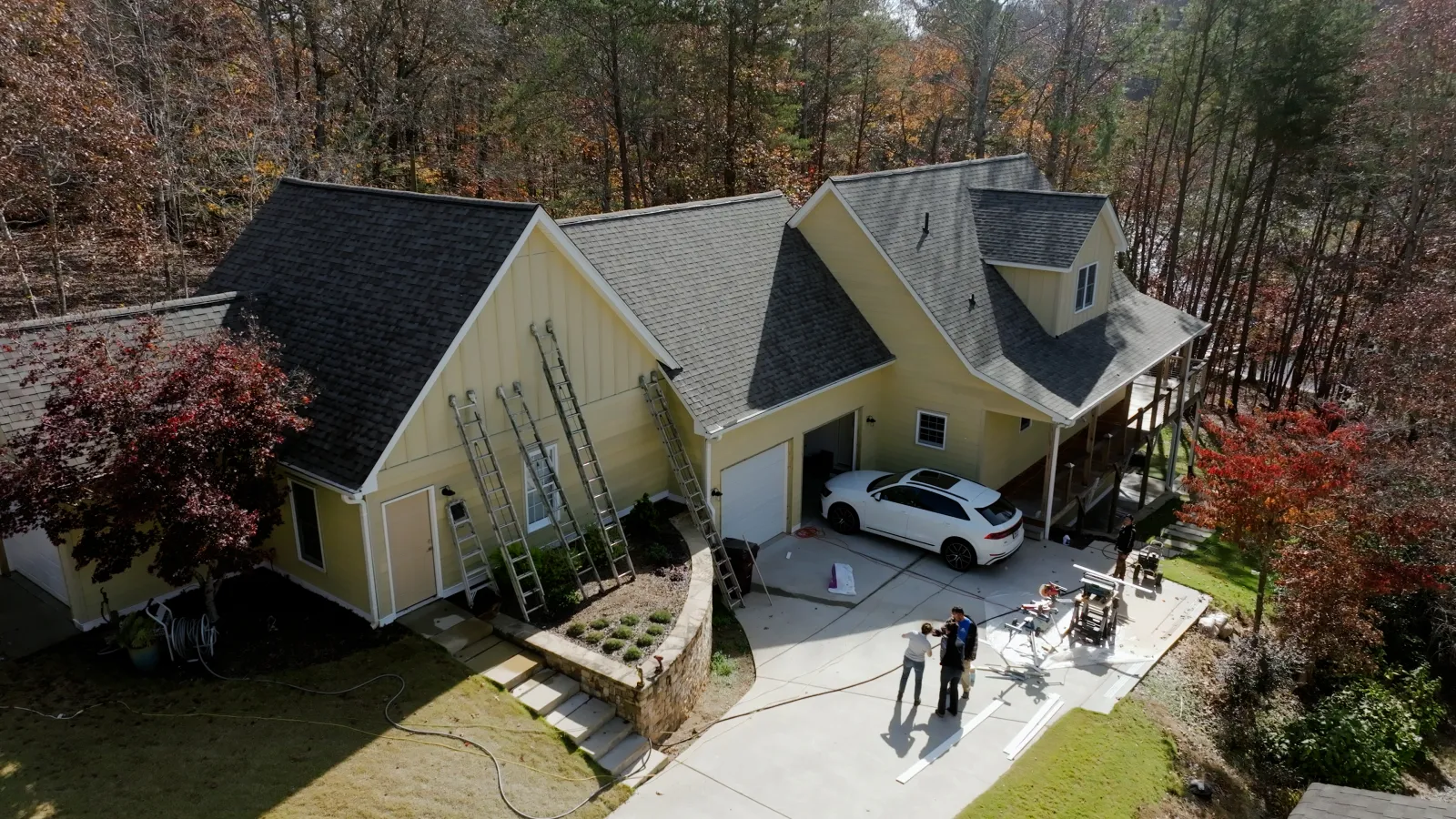 A yellow house with a white car parked in the driveway and people standing near outdoor equipment on a sunny day.