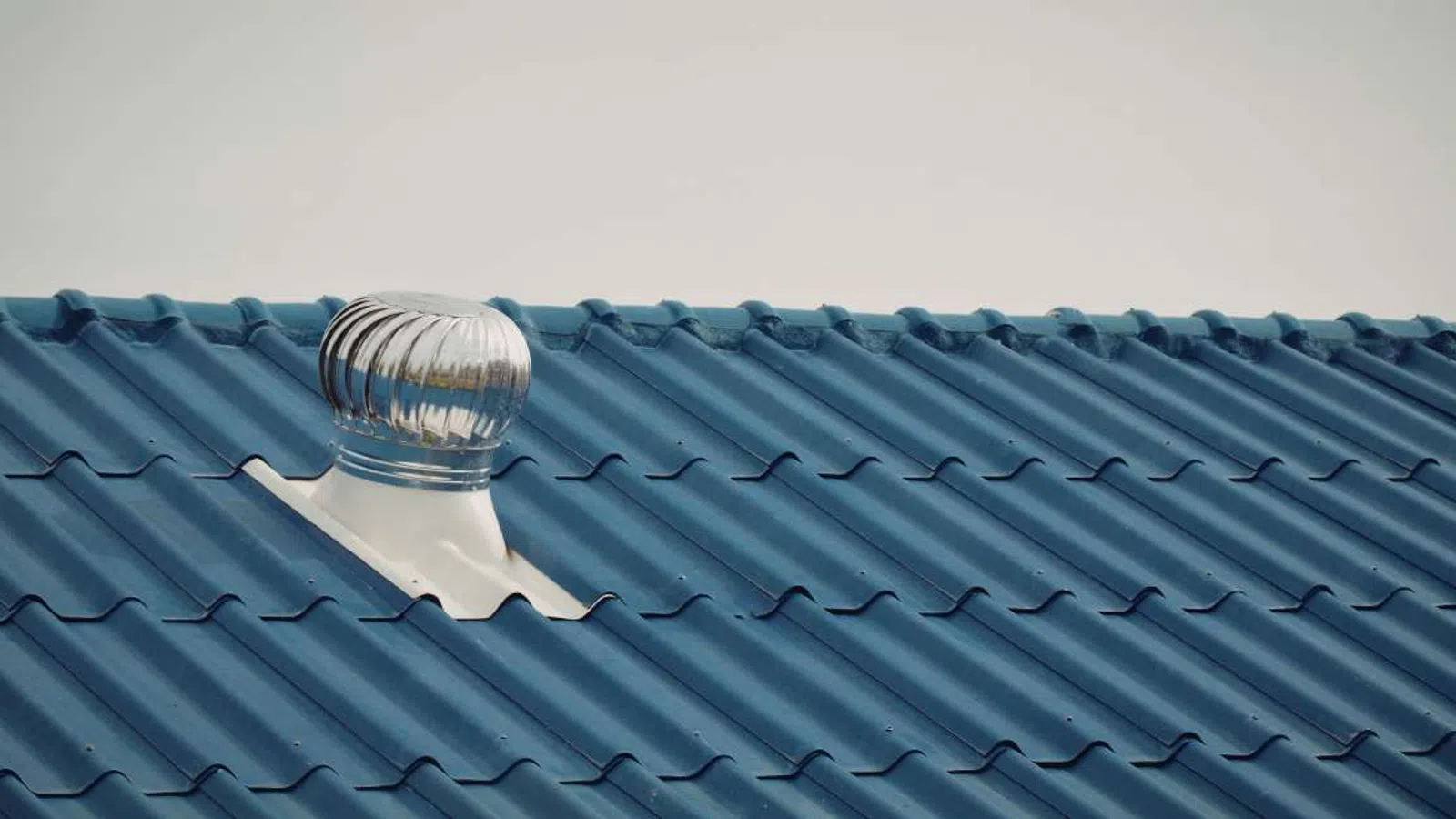 Silver metal turbine roof vent on blue corrugated metal roofing under clear sky.