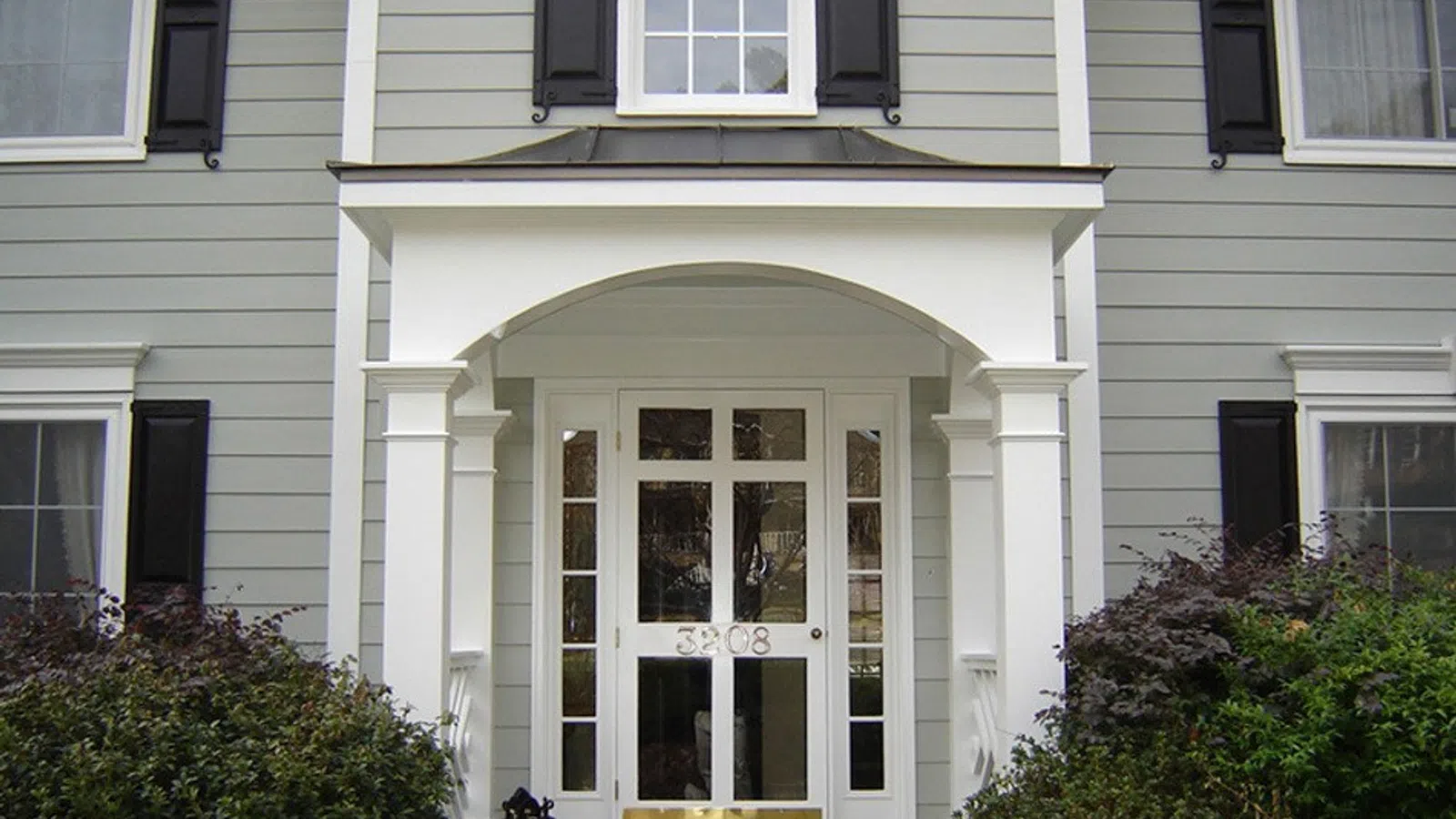 Elegant home entrance with a white portico, black shutters, and lush greenery framing the doorway.