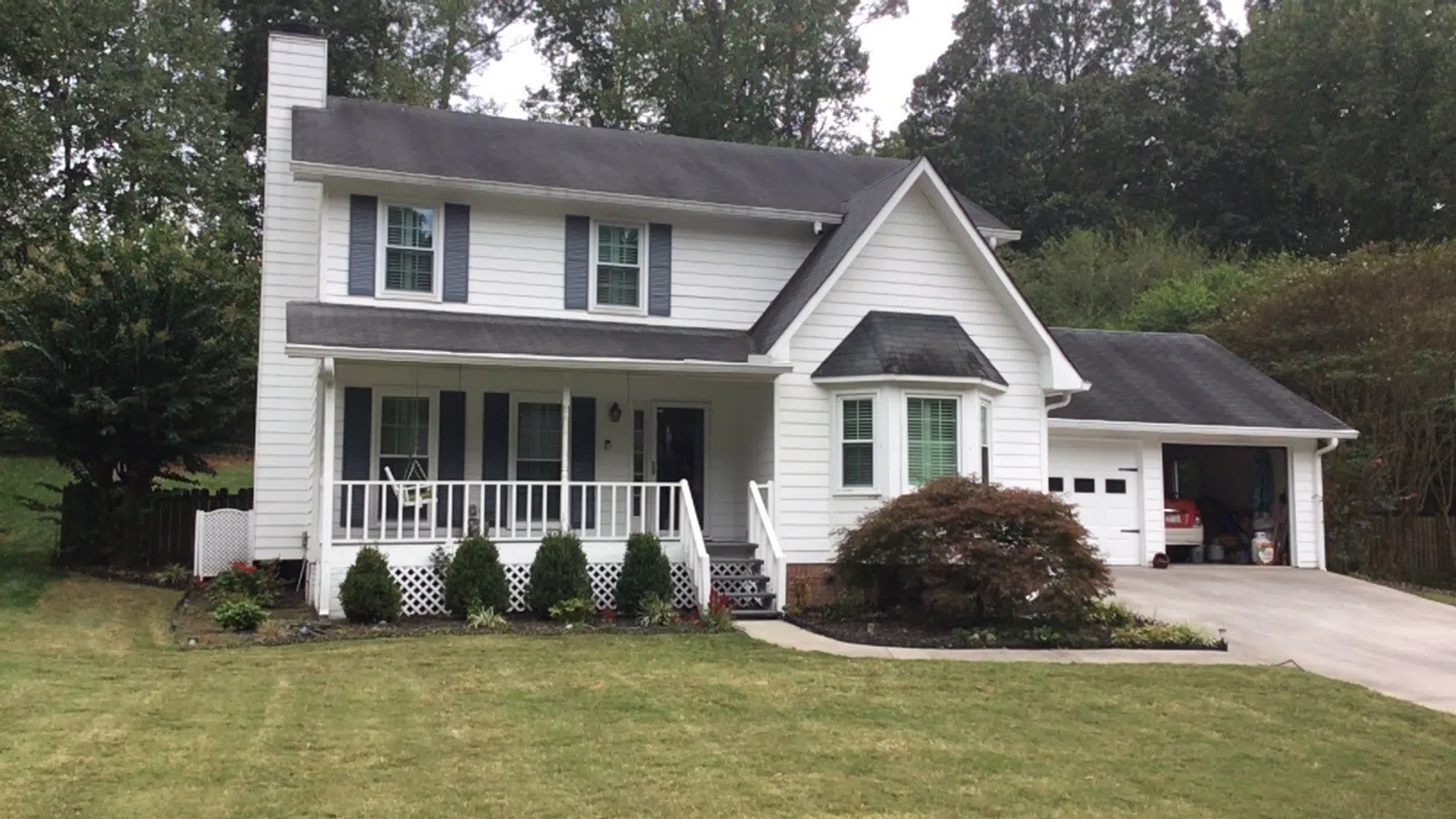 Charming two-story white house with a porch, green lawn, and garage surrounded by trees.