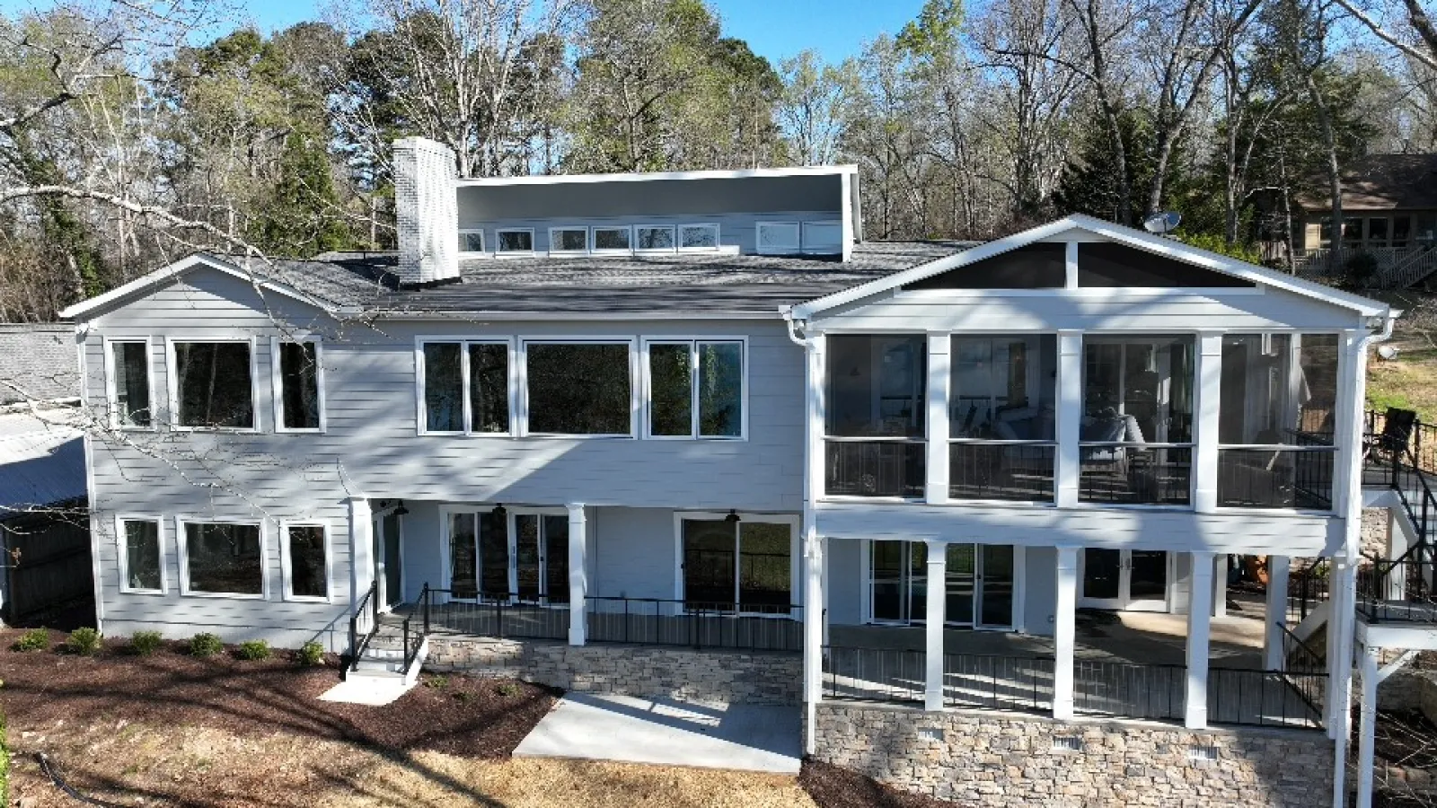 Modern two-story gray house with large windows, screened porch, stone foundation, and surrounding trees in early spring.