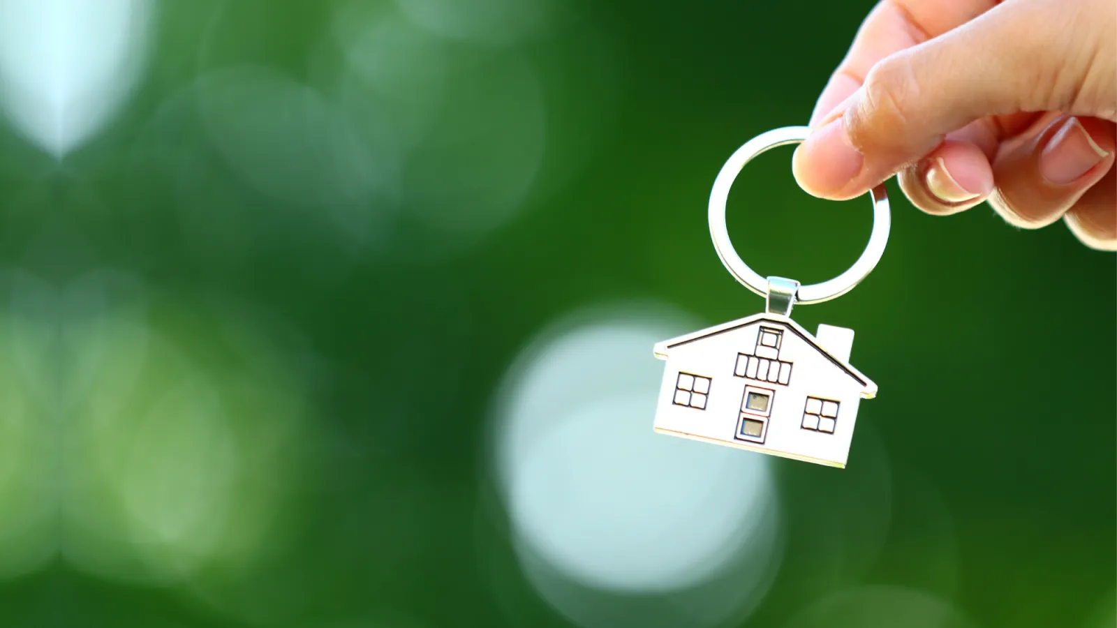 Hand holding house-shaped keychain against a blurred green background symbolizing home ownership.