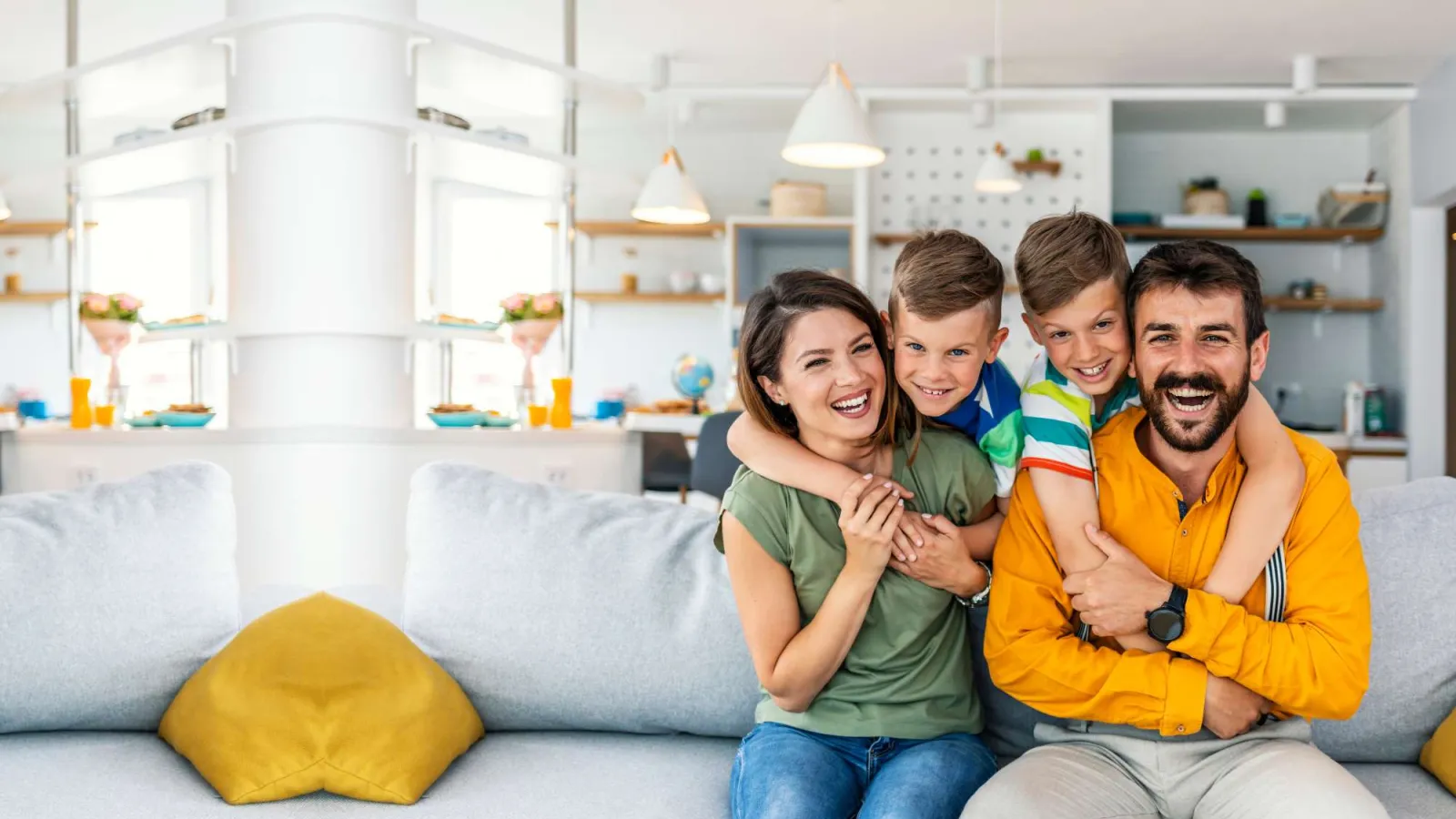 Happy family of four sitting on a gray couch in a bright modern living room with yellow pillows.
