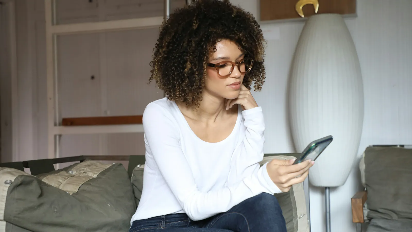 Woman with curly hair and glasses sitting on couch using smartphone in cozy living room setting