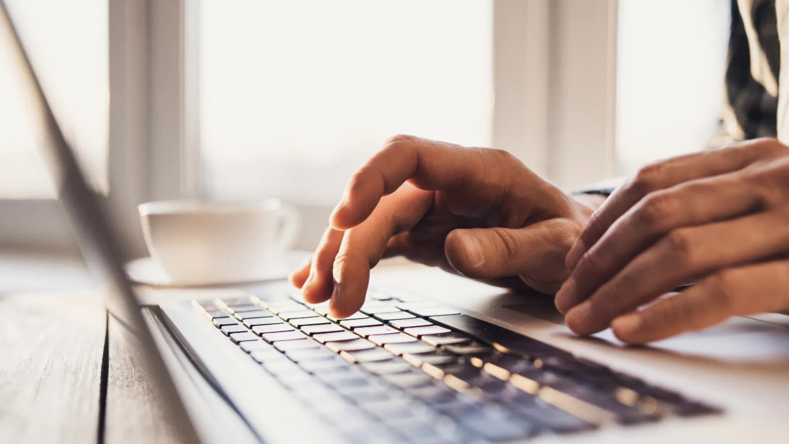 Close-up of hands typing on a laptop keyboard with a blurred coffee cup and window in background