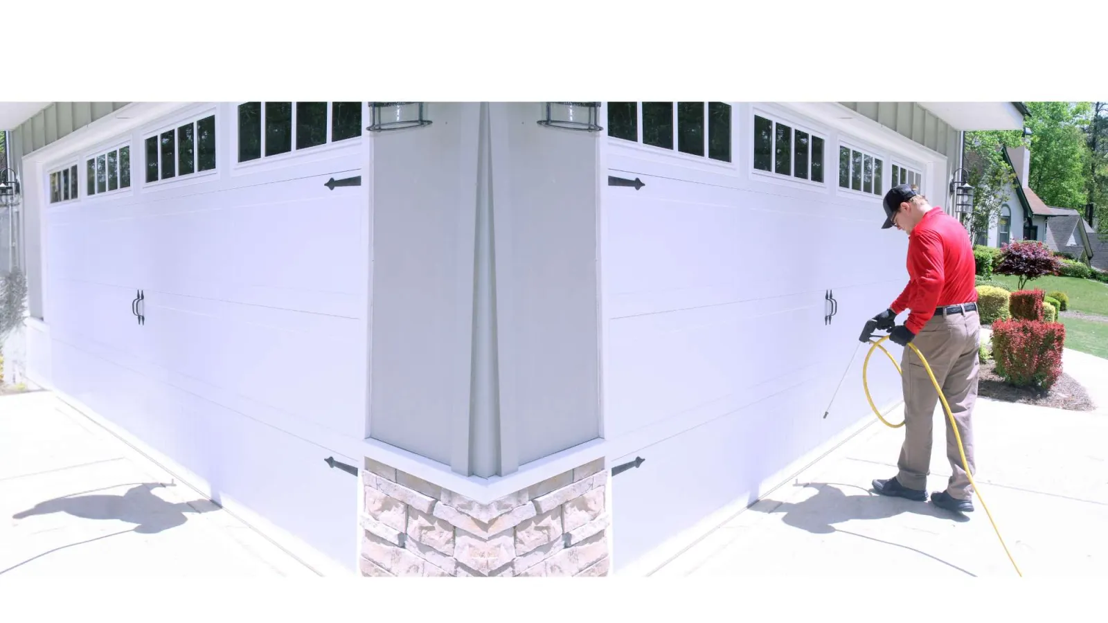 Man in red shirt power washing a white garage door on a sunny day in a suburban neighborhood