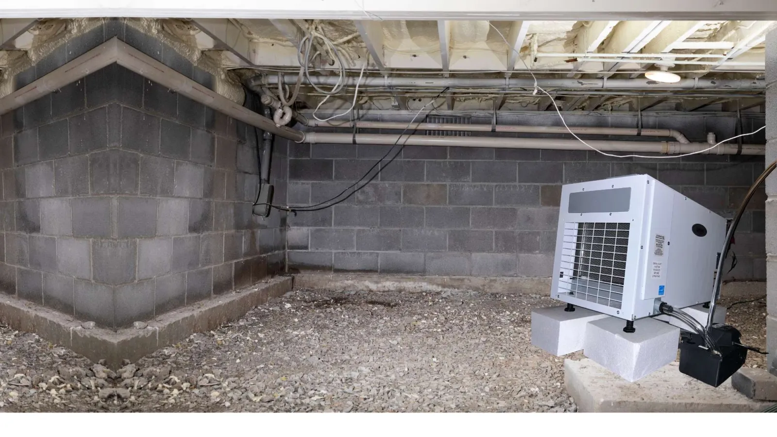 crawl space Dehumidifier installed on foam blocks in a dry, gravel-covered crawl space with concrete block walls and exposed ceiling pipes.