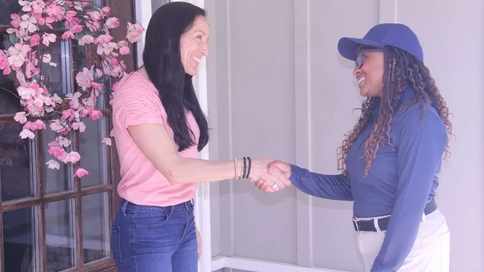 Two women smiling and shaking hands at a doorway decorated with pink flowers, greeting each other warmly