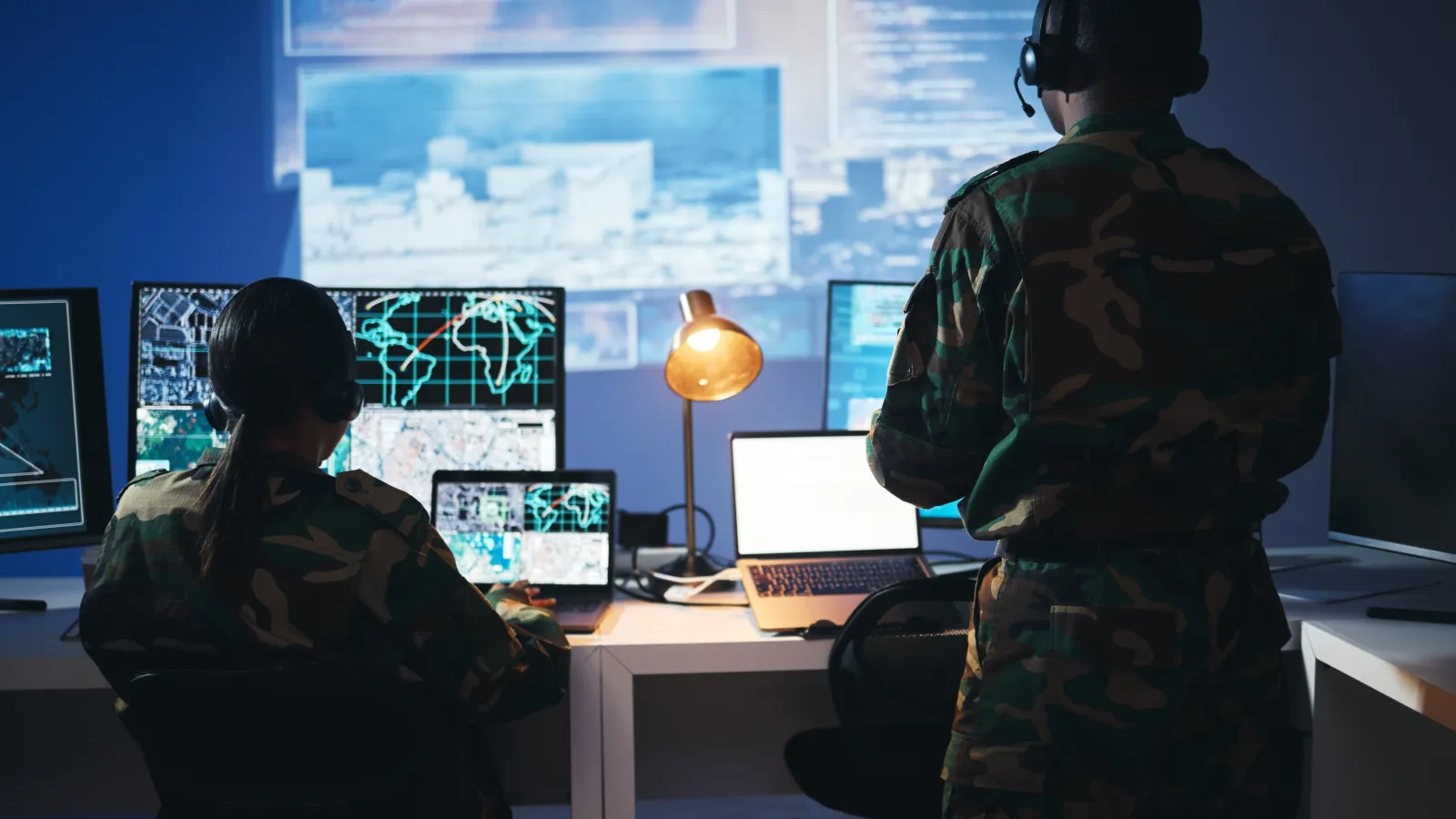 Military personnel monitoring surveillance data on multiple computer screens in a dark command center.