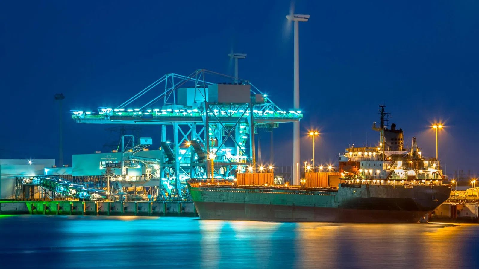 Cargo ship docked at illuminated port terminal with cranes and containers during nighttime.