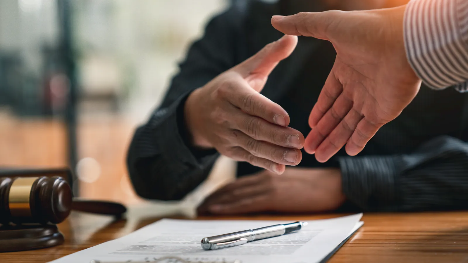 Two people reaching out to shake hands over a legal contract with a gavel on the table, symbolizing agreement.