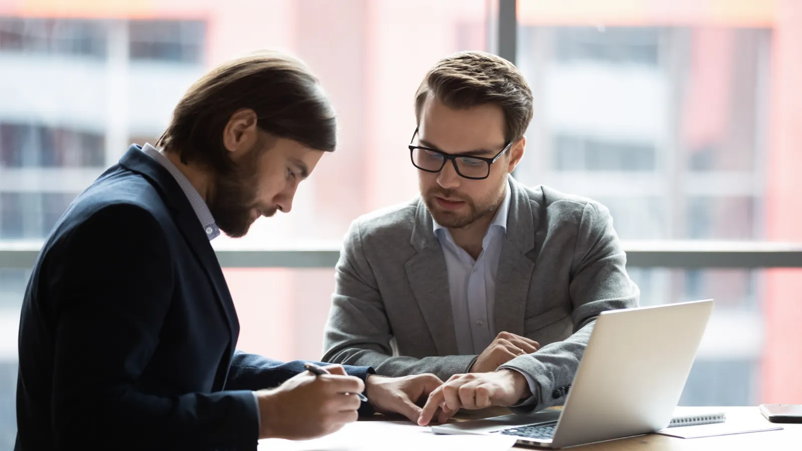 Two businessmen reviewing documents together at a desk with a laptop in a modern office setting.