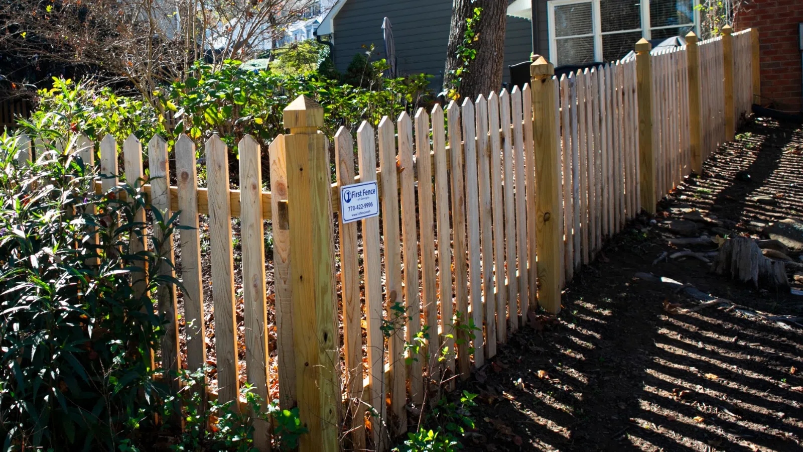 A well-maintained wooden fence surrounding a garden, with greenery and shadows cast by sunlight.