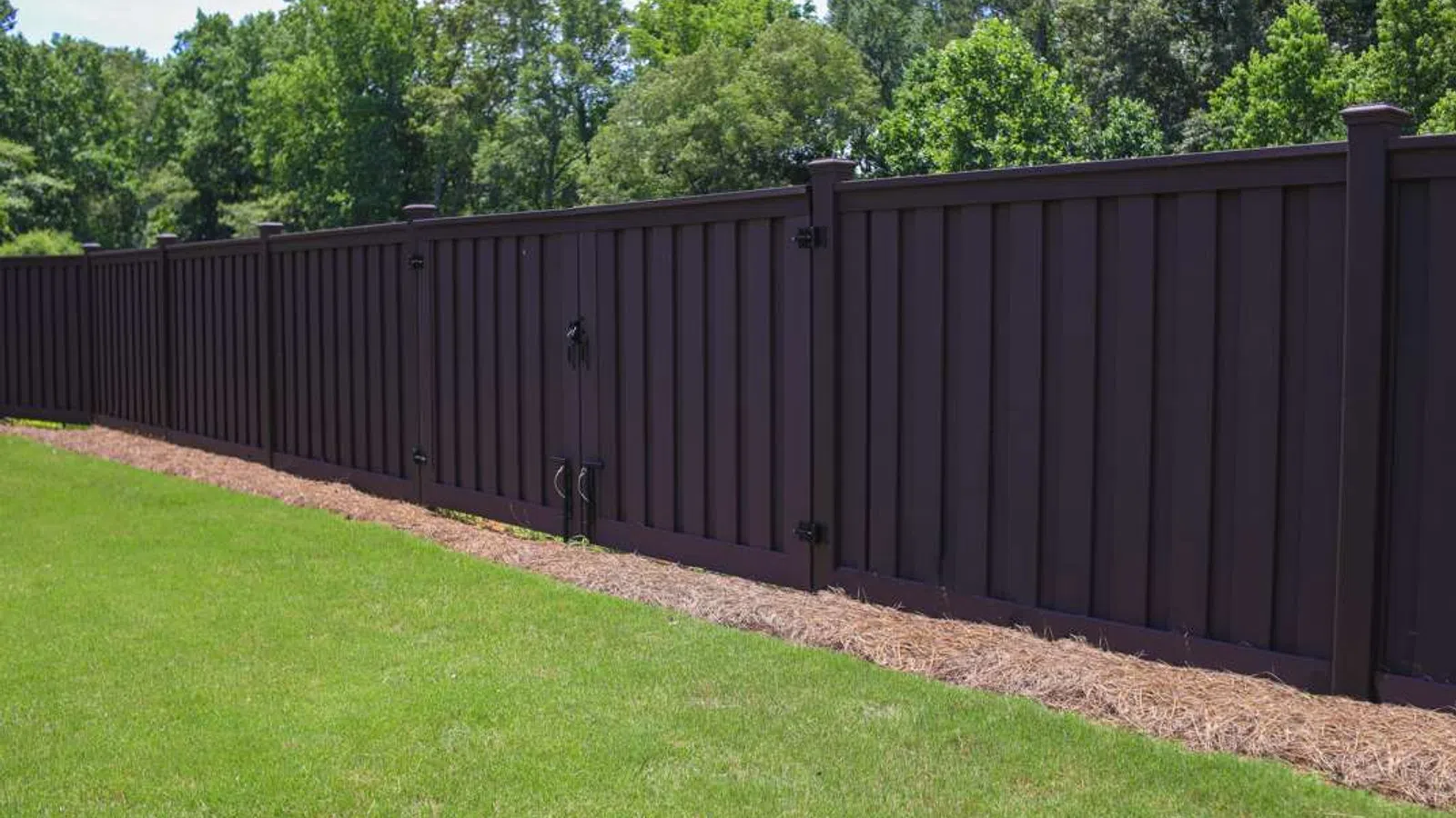 Long dark brown wooden fence with gate enclosing a grassy yard next to green trees in sunlight