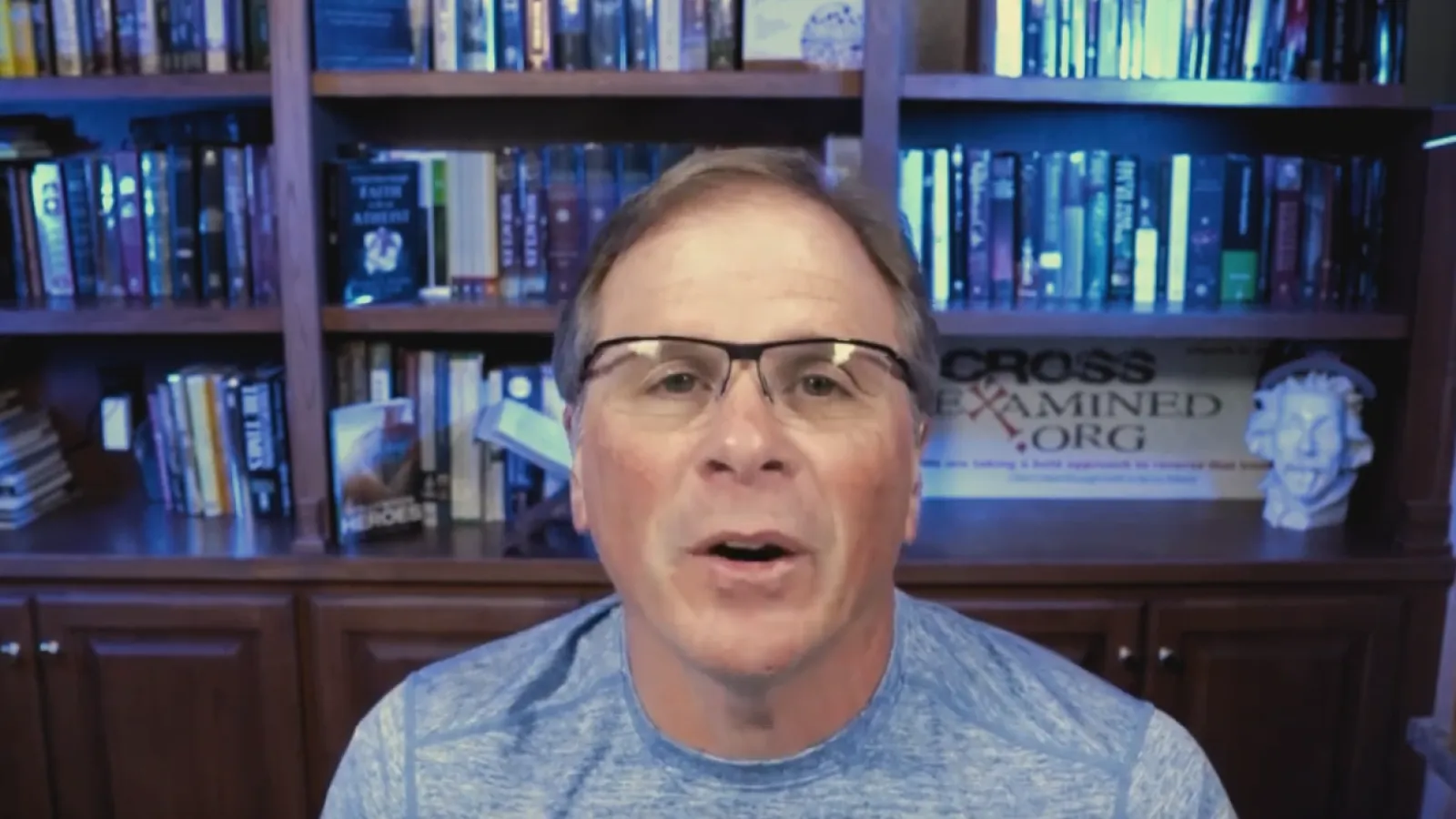 Man wearing glasses and blue shirt speaking in front of a bookshelf filled with books and decorations.