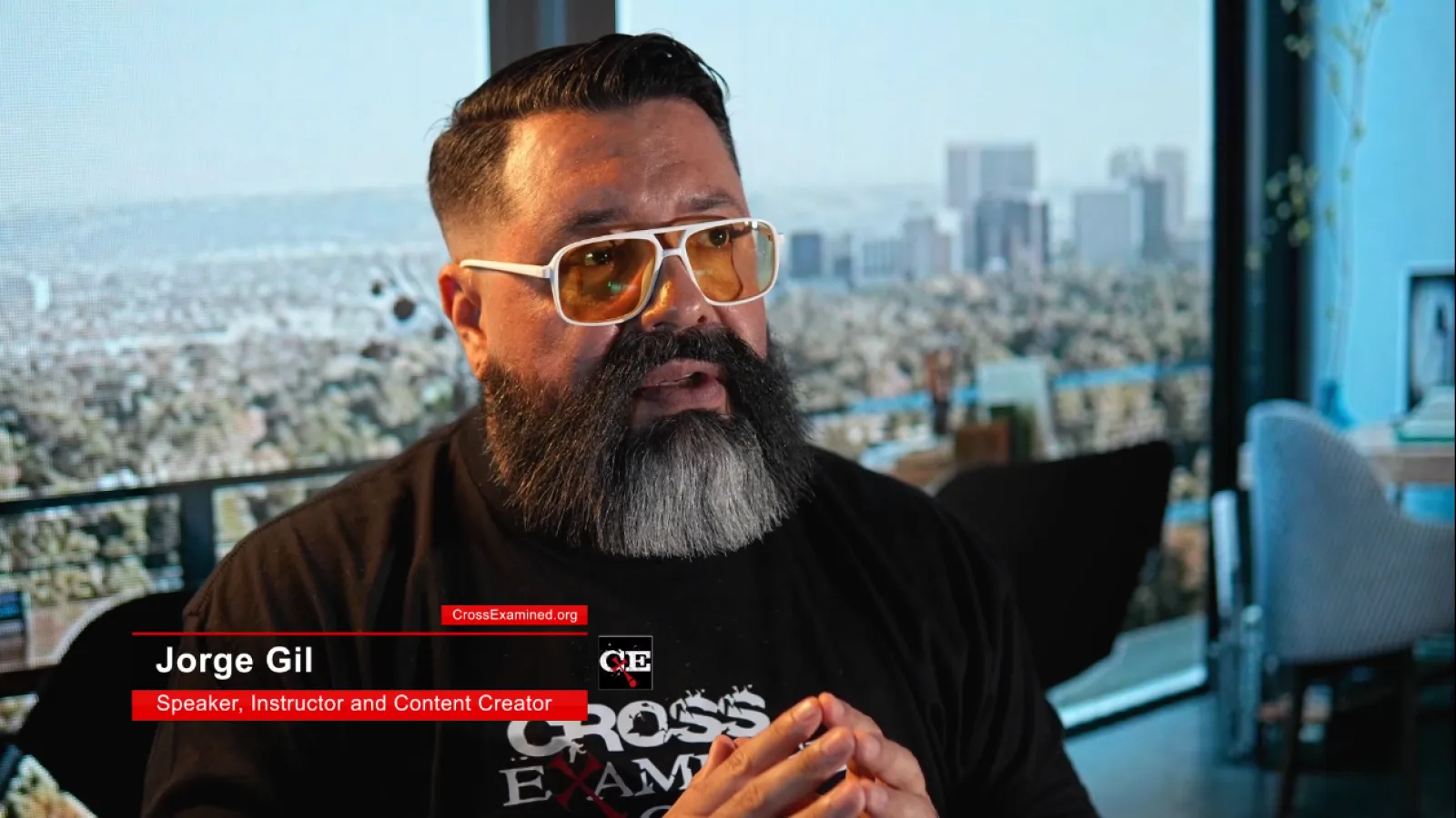 Man with beard and glasses speaking indoors with a cityscape view through large windows behind him