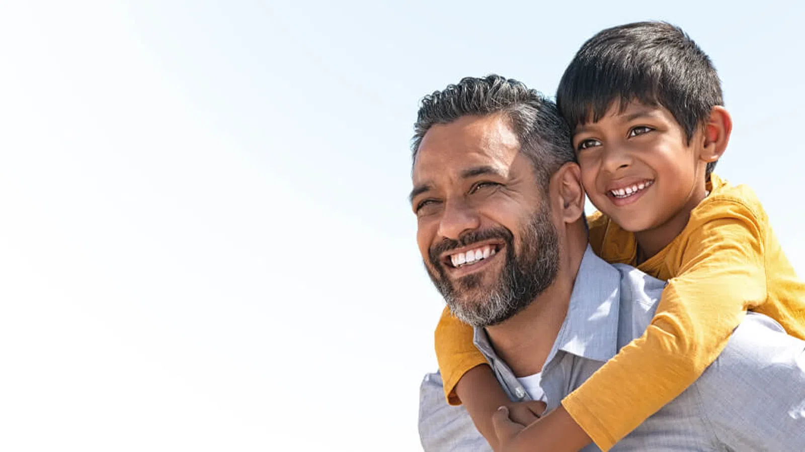 Smiling father giving piggyback ride to happy son against a clear sky background in daylight.